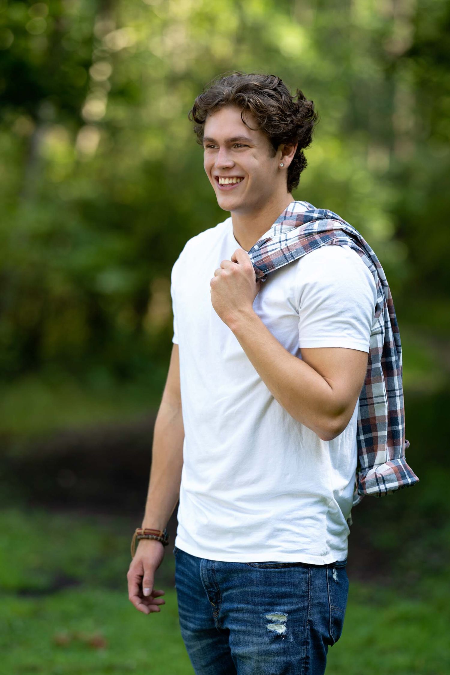 Person in white t-shirt and jeans stands casually outdoors with plaid shirt draped over shoulder in natural setting.