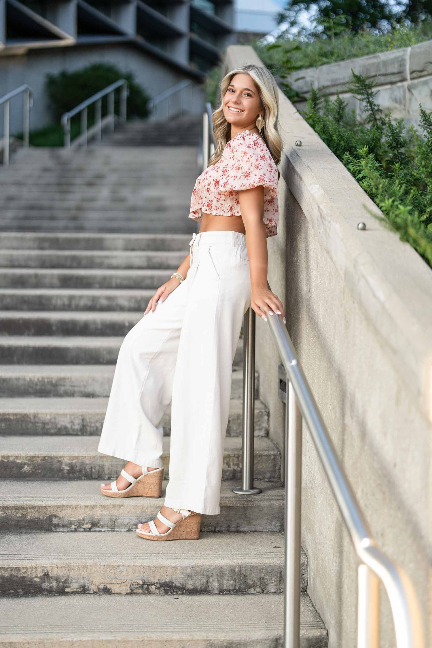 A woman in a floral top and white pants poses on concrete steps in an outdoor setting.