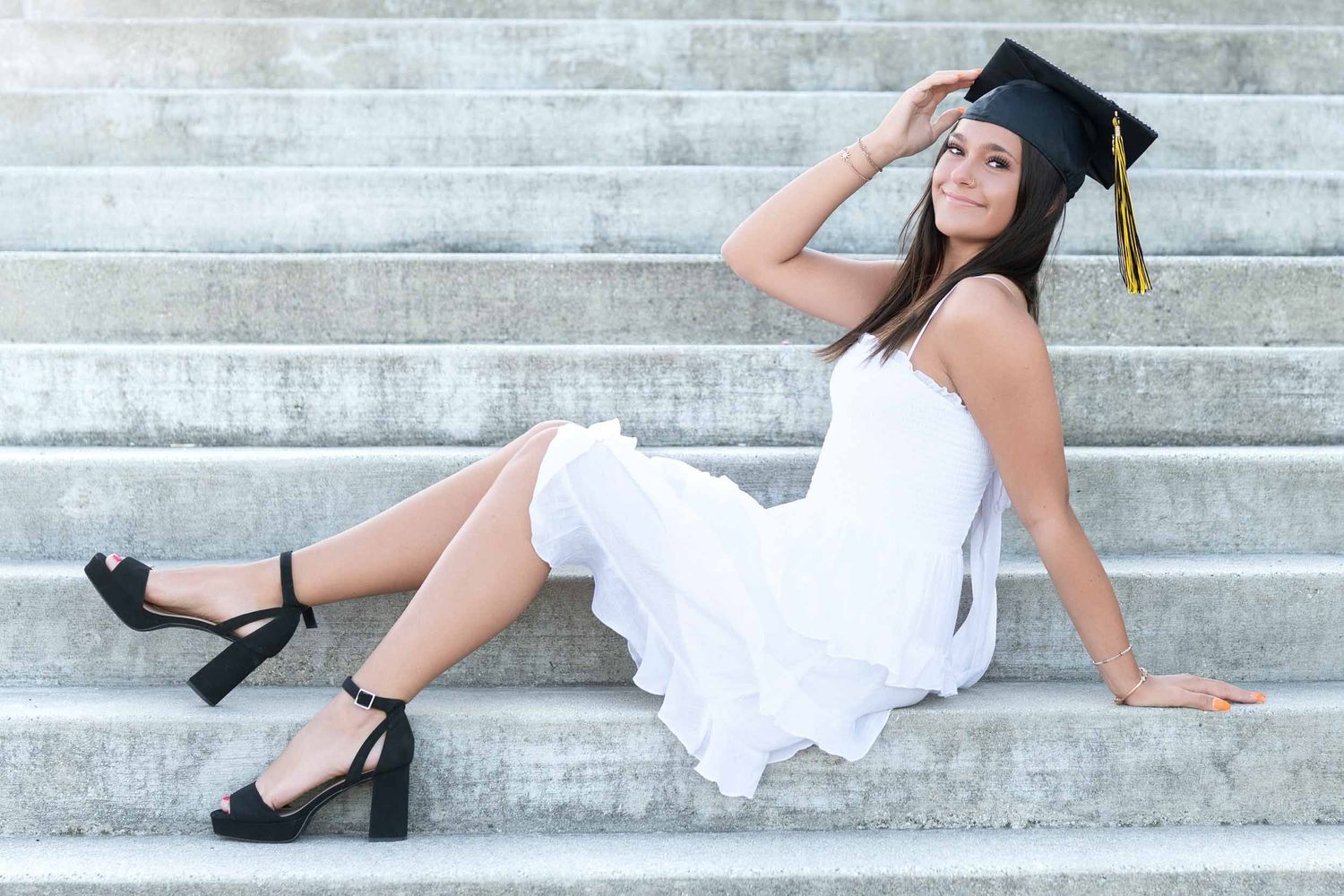 Graduate in white dress and cap sits elegantly on concrete steps while posing during outdoor graduation photoshoot.