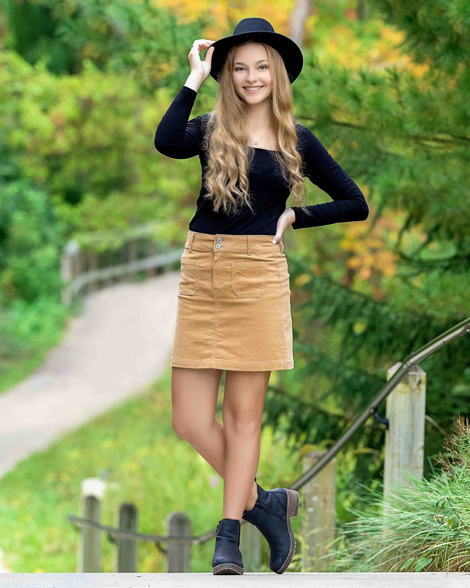 Young woman in black top, tan mini skirt, and black hat poses on a fence in a lush green park setting.