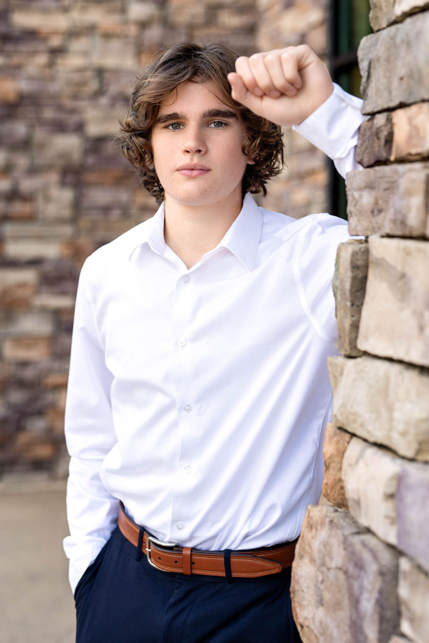 A person in a crisp white button-down shirt and navy pants poses against a stone wall backdrop.