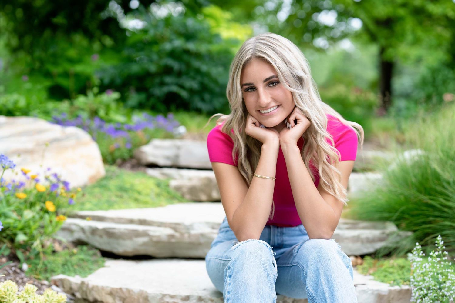 Person in casual attire sits on stone steps surrounded by lush greenery and wildflowers in a garden setting.