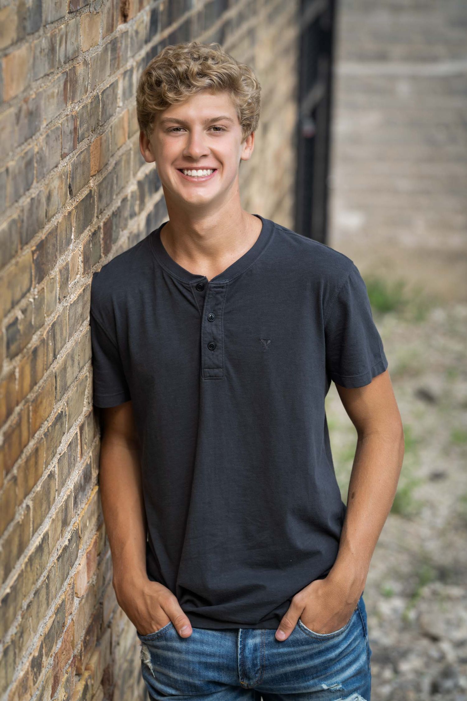 A person in a black henley shirt and jeans poses casually against a brick wall in an outdoor portrait series.