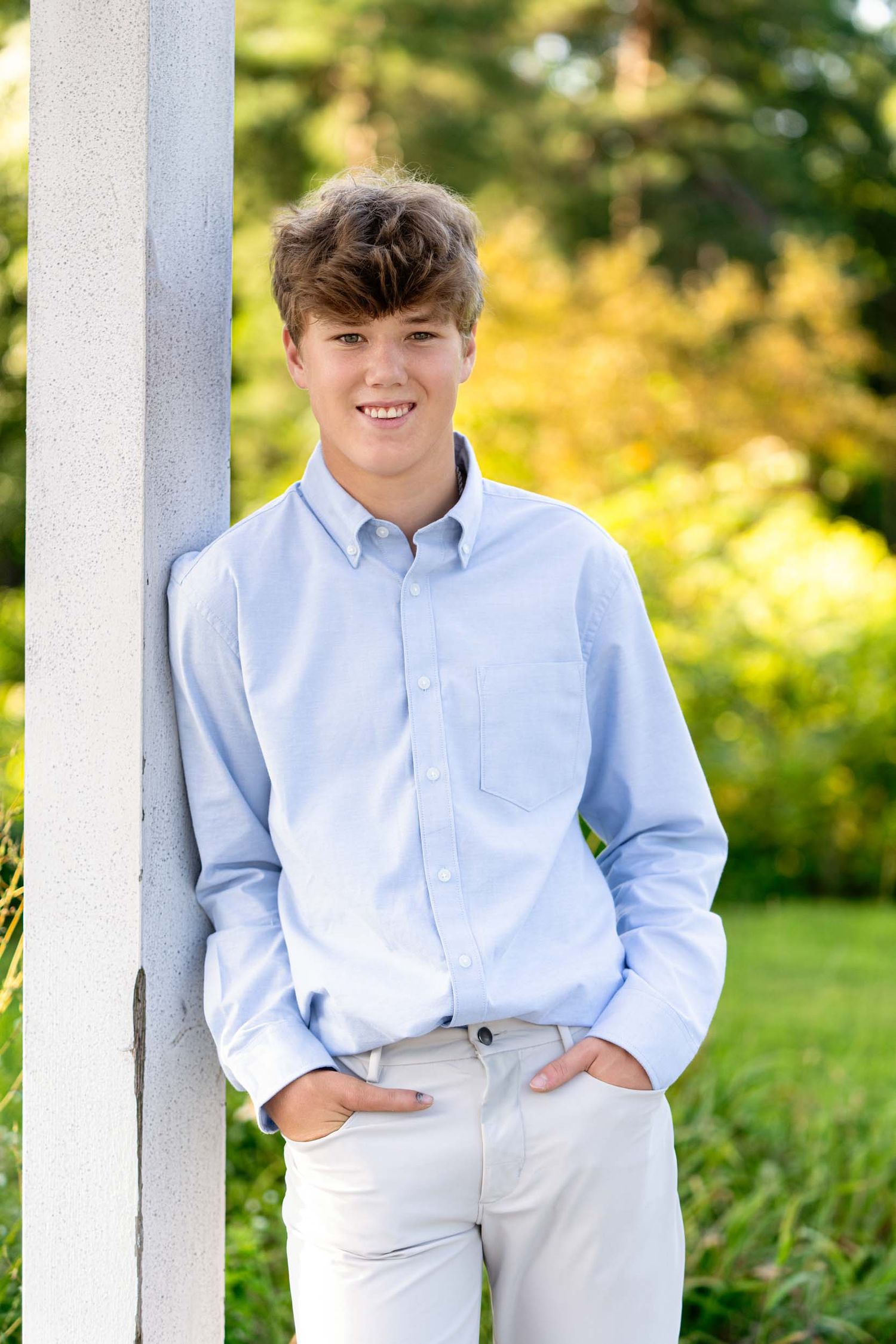 A young person in a light blue button-down shirt and white pants poses against a white column outdoors.