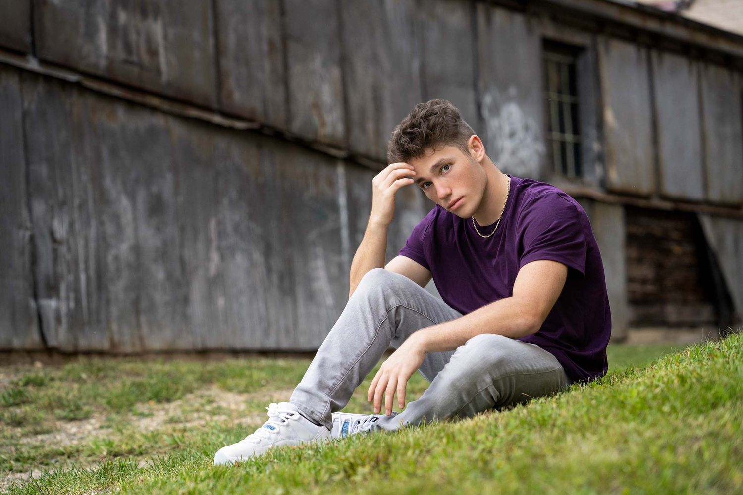 Person in purple shirt and gray pants relaxing on grassy slope next to rustic wooden wall in outdoor setting.