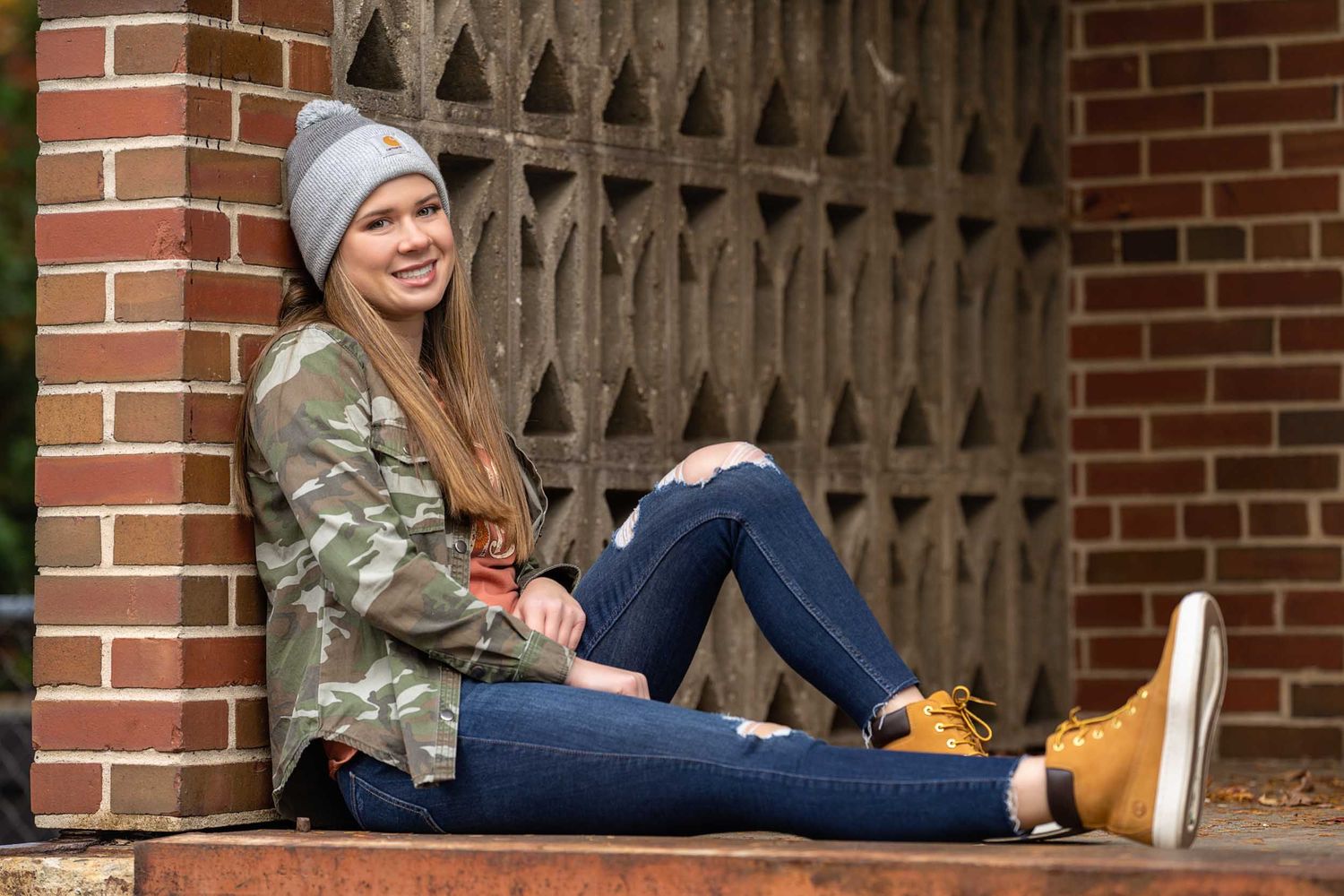 Someone in a camo jacket and beanie sits casually against a brick wall with decorative gothic architecture.