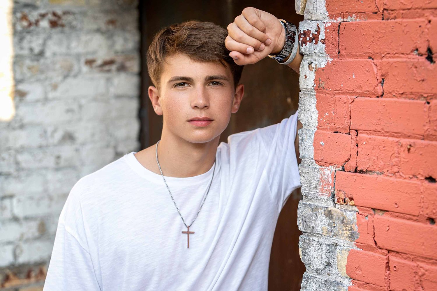 Person in white t-shirt poses against red brick wall background with cross necklace.