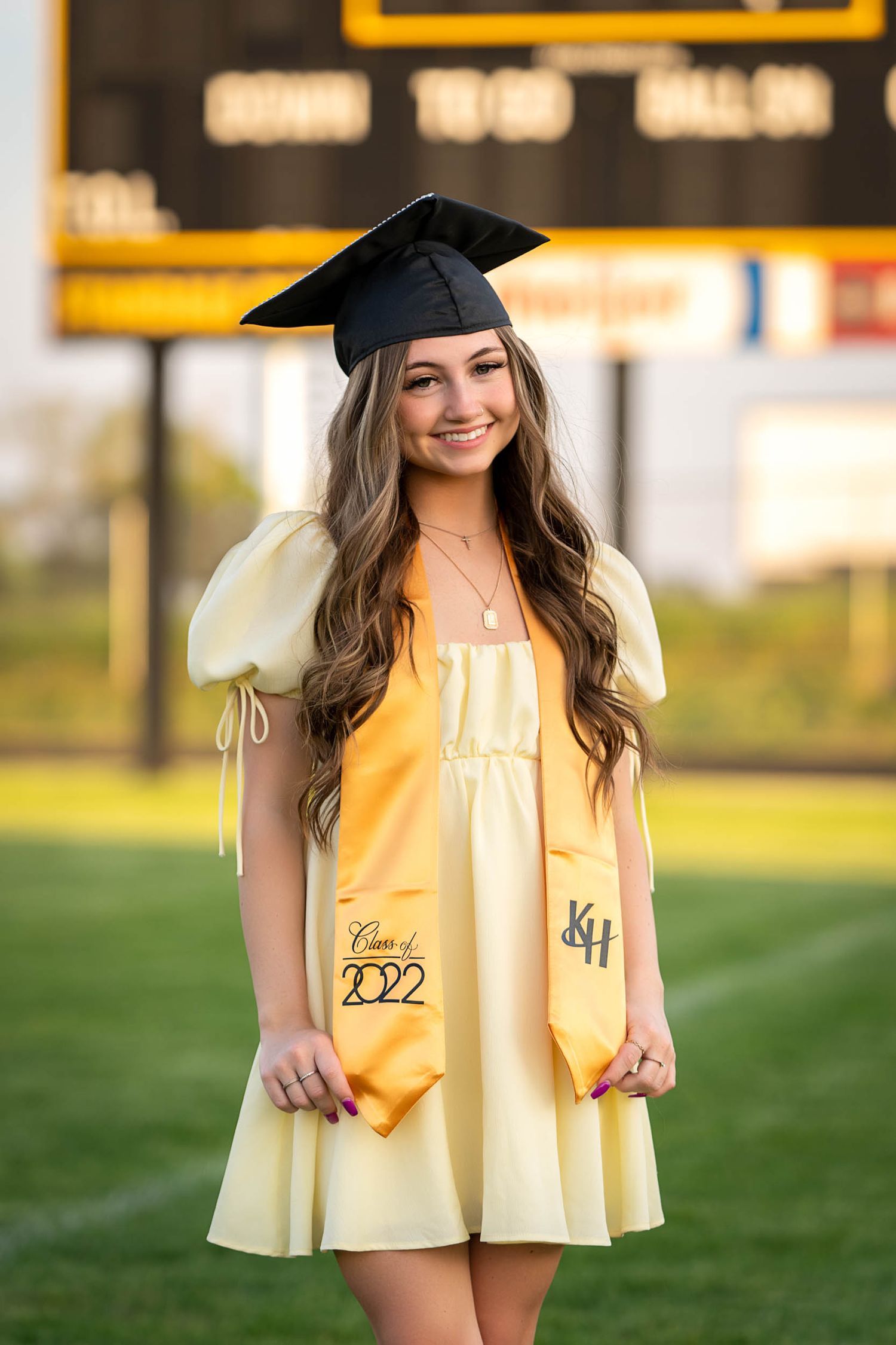 A graduate wearing a yellow dress and graduation cap poses outdoors on a sunny day with a Class of 2022 sash.