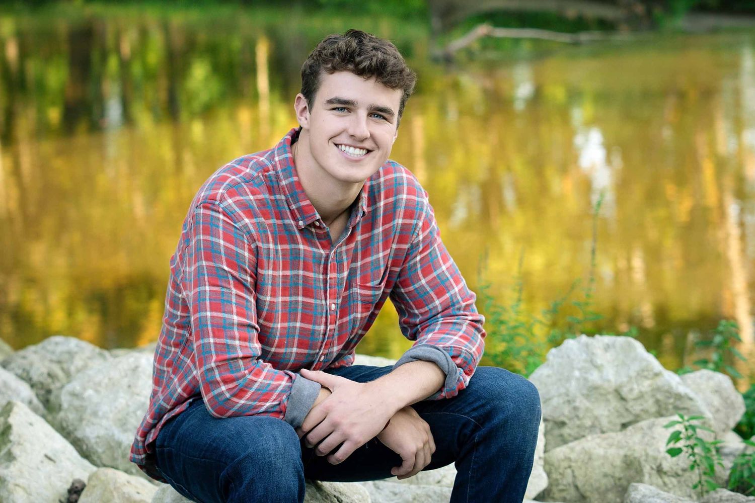 A person in a red plaid shirt sits on large rocks outdoors during golden hour with autumn foliage in the background.