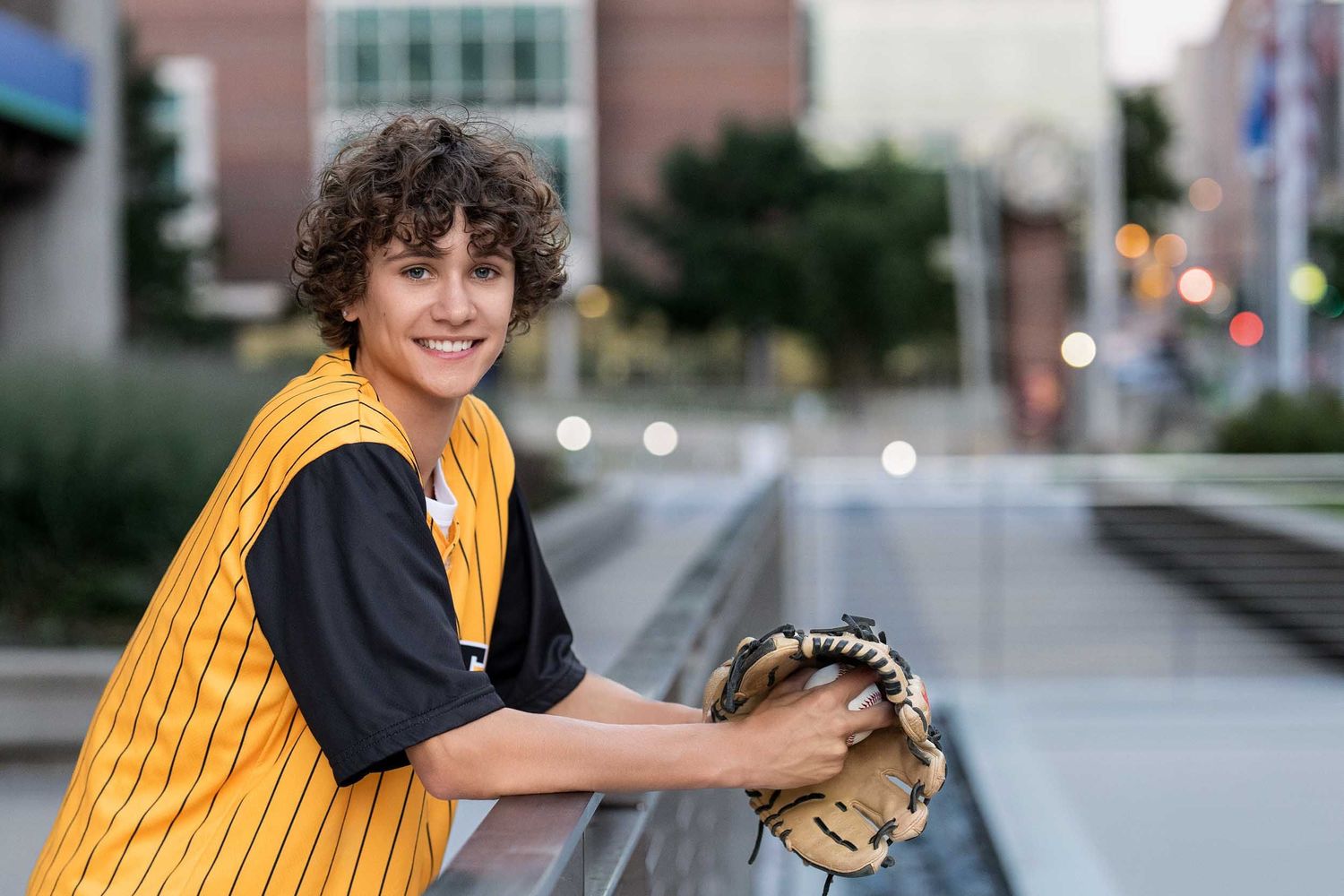 A person in a yellow striped dress and black bow holds a baseball glove while standing in an urban setting.