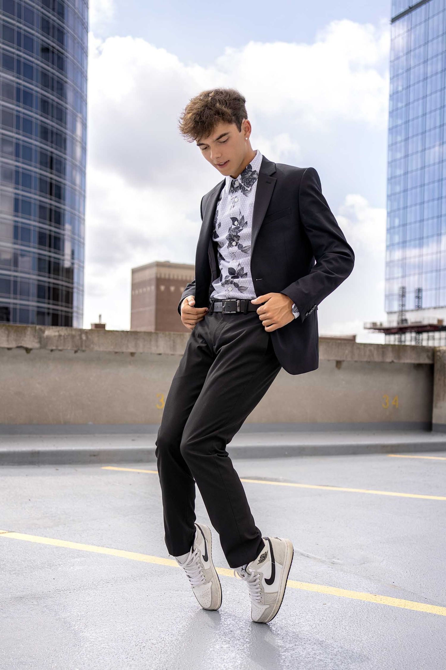 Stylish black suit and sneakers outfit photographed on urban rooftop with modern glass buildings in background.