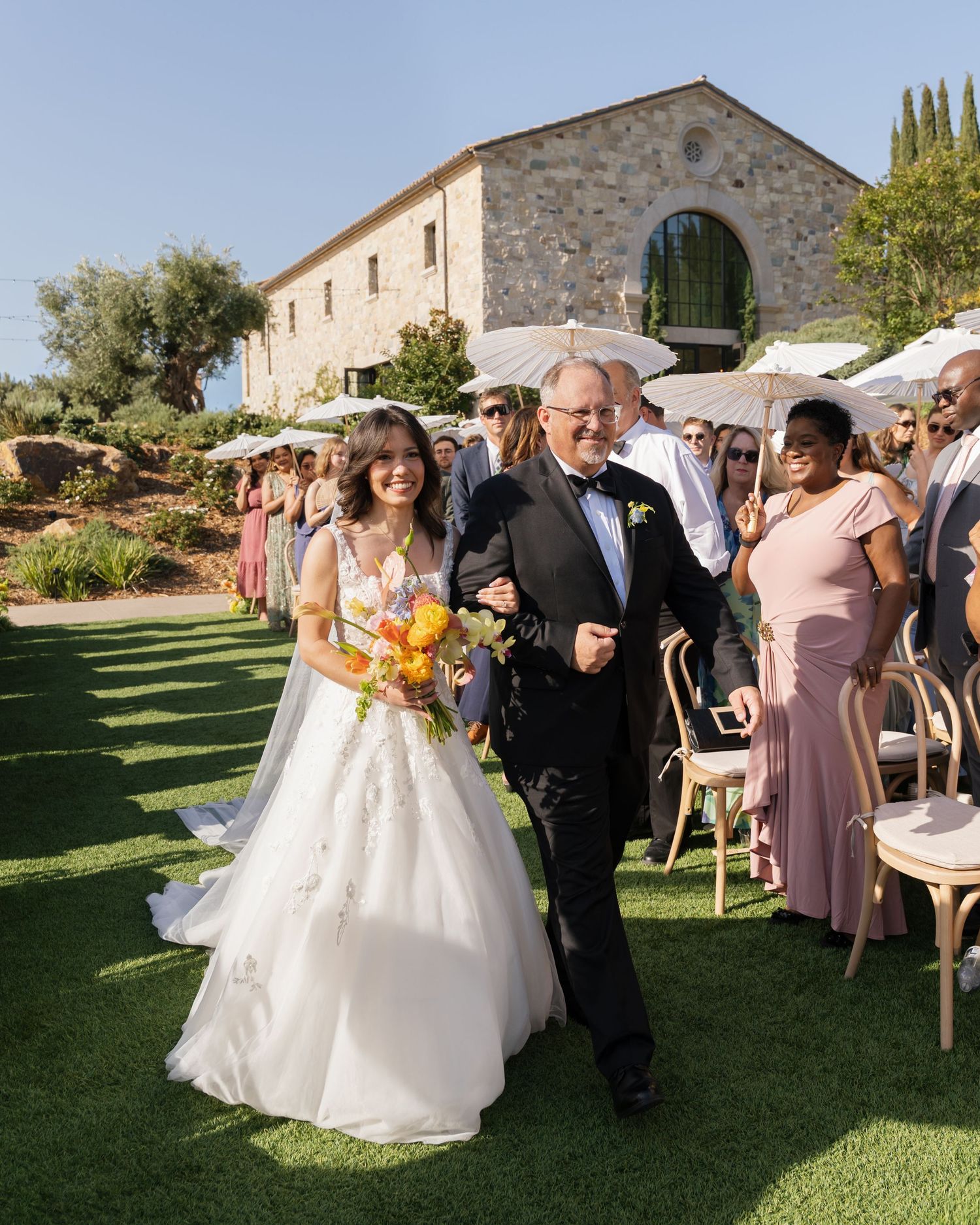 Wedding guests gather on a lush green lawn outside a rustic stone building during an outdoor celebration.