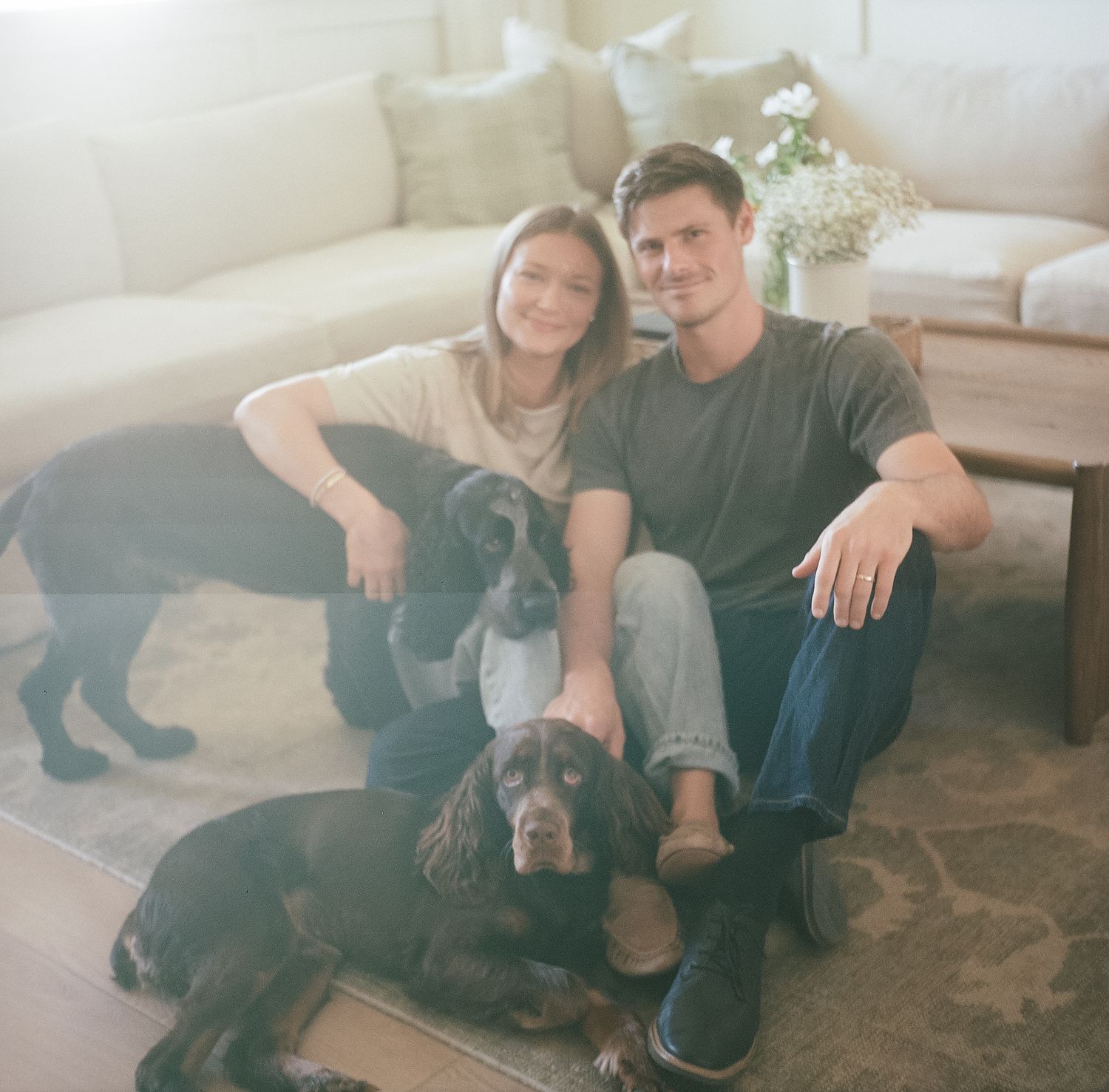 A smiling couple sits on the floor with two dark-colored dogs in a bright, cozy living room.