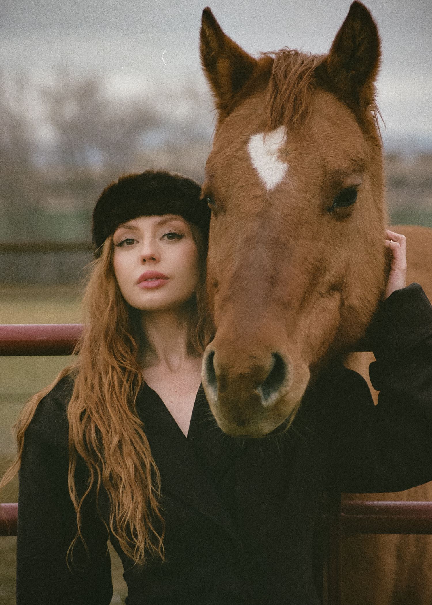Young woman in black outfit and fur headband poses beside a chestnut horse with a white star marking outdoors.