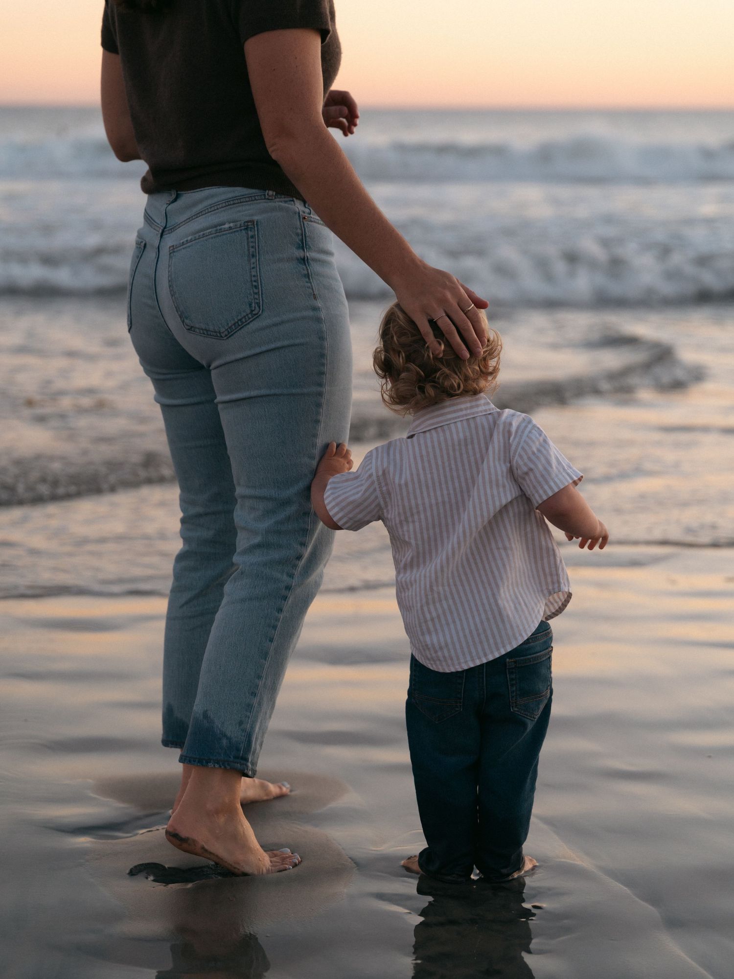 Parent and child stand barefoot at the beach shore during sunset as gentle waves lap at their feet.