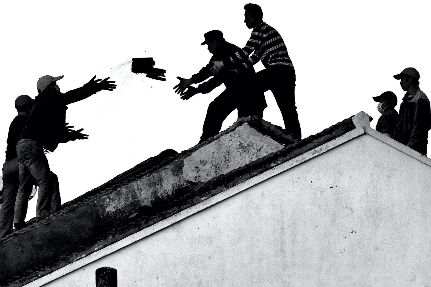 Silhouetted figures pass bricks along a rooftop line in a dramatic black and white construction scene.