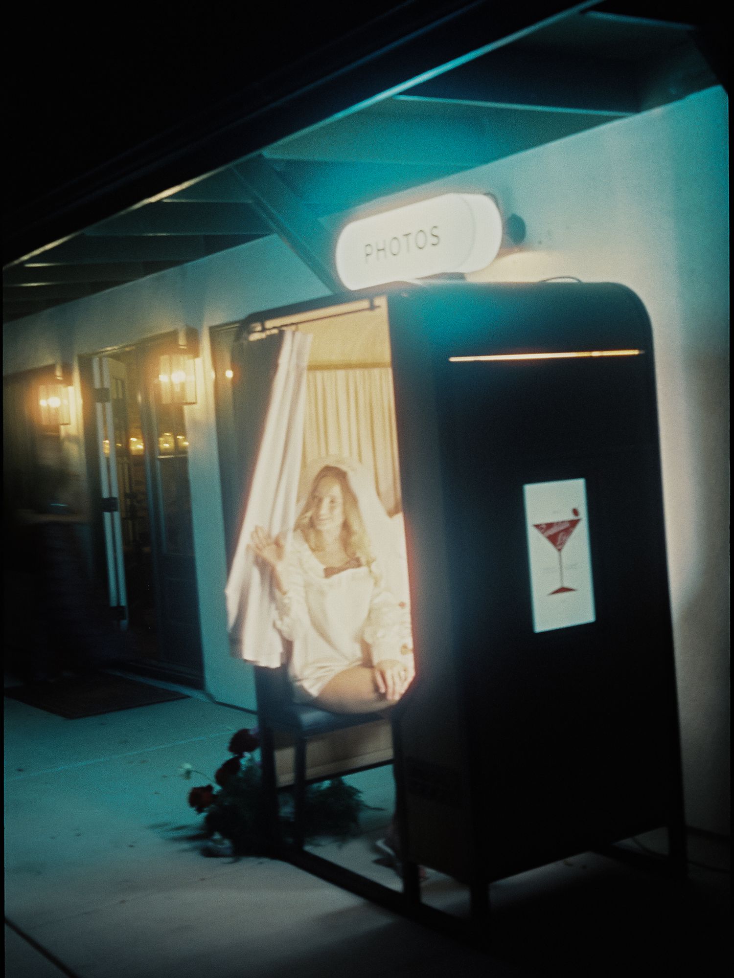 Woman sitting inside a vintage photo booth with glowing PHOTOS sign and martini glass graphic at night.