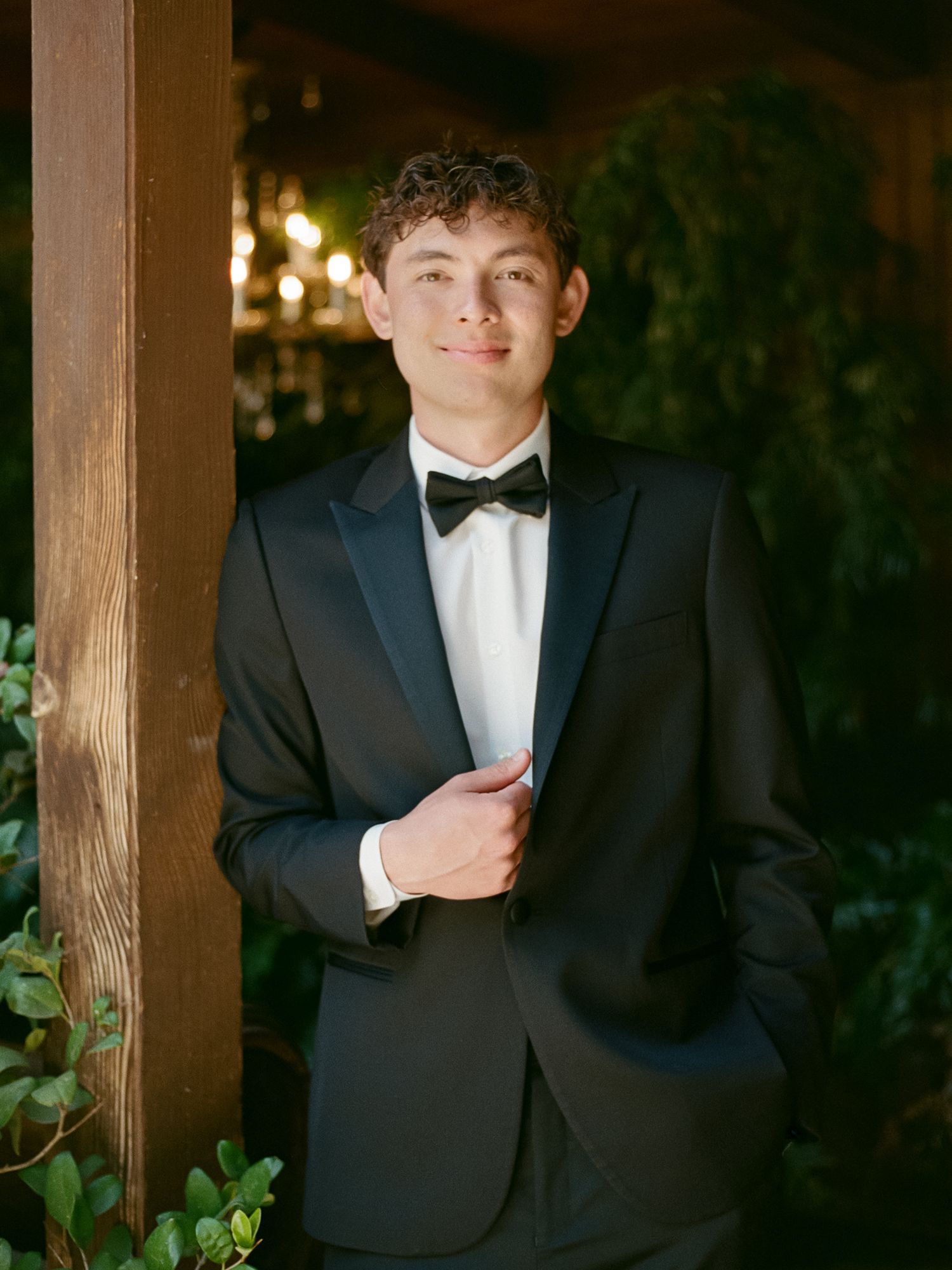Young man in black tuxedo with bow tie posing confidently outdoors near wooden post with greenery.
