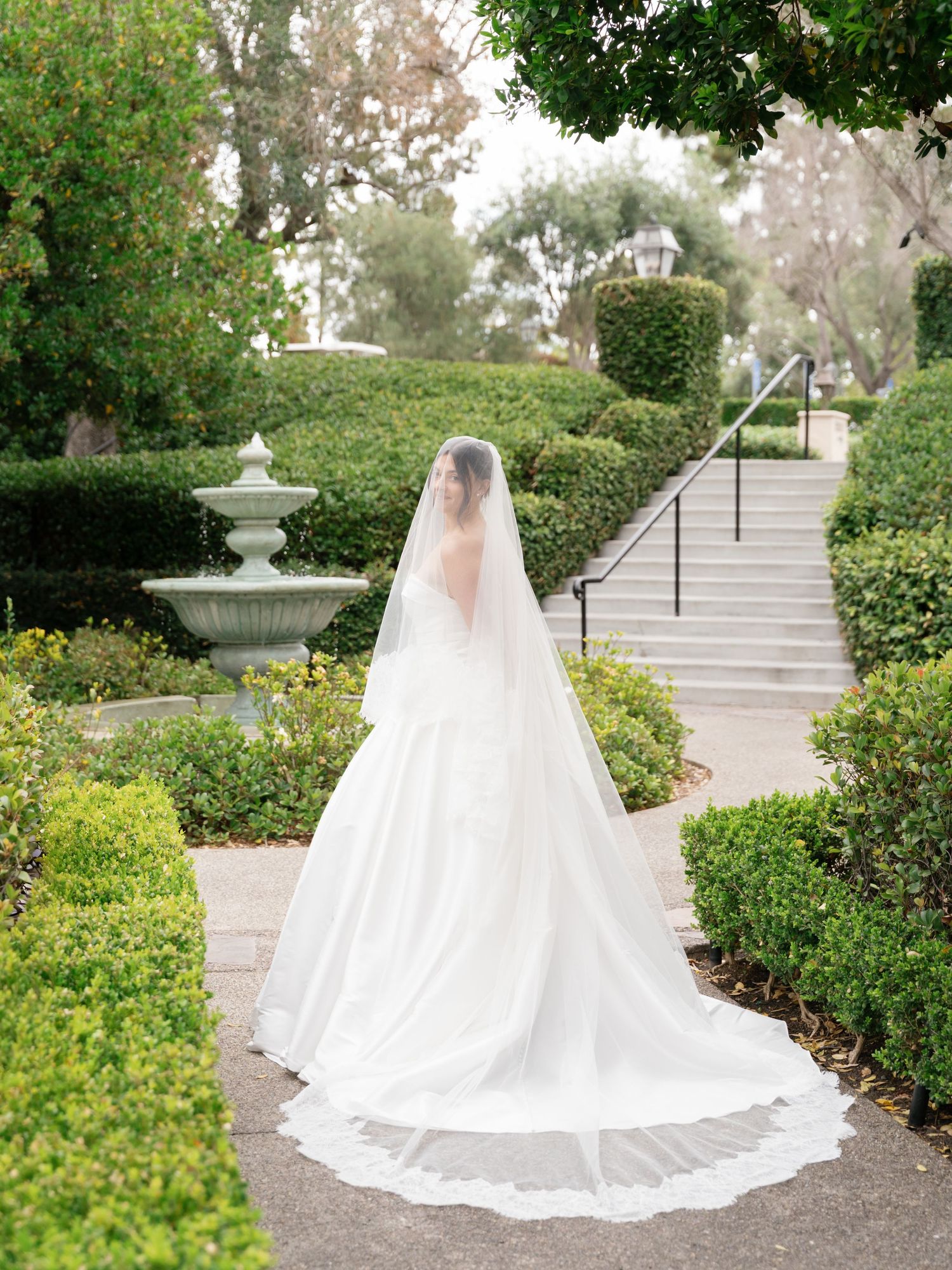 Bride in elegant white gown with long veil stands in lush garden with fountain and manicured hedges.