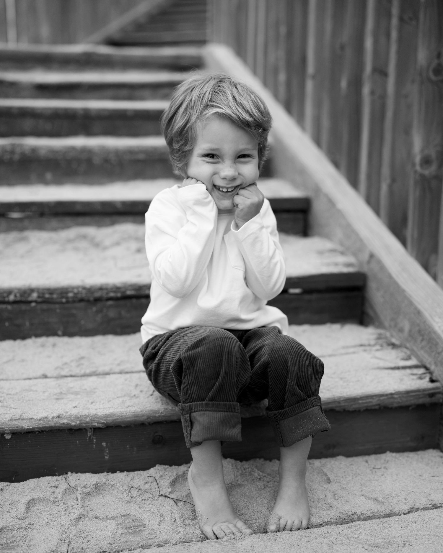 Smiling barefoot child sitting on wooden steps in black and white portrait photo.