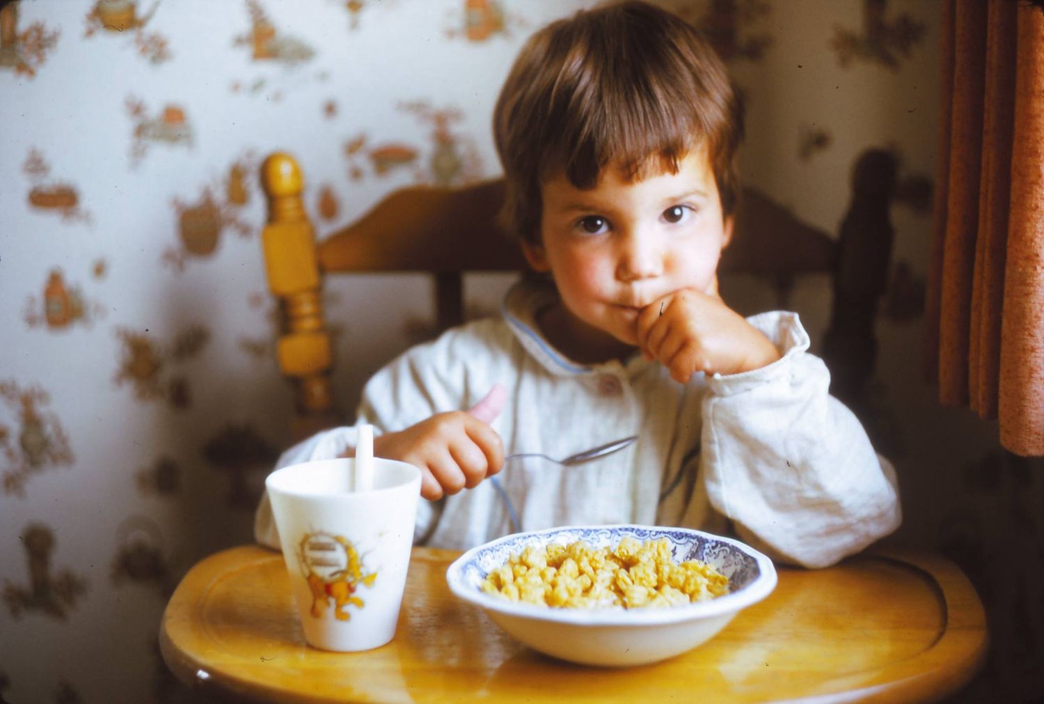 Young person in white sitting at table with bowl of cereal against floral wallpaper background.
