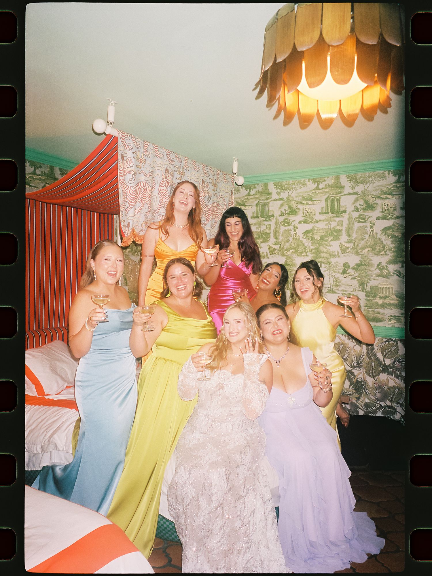 Bride in white gown poses with bridesmaids in colorful dresses, holding champagne in a decorated room.