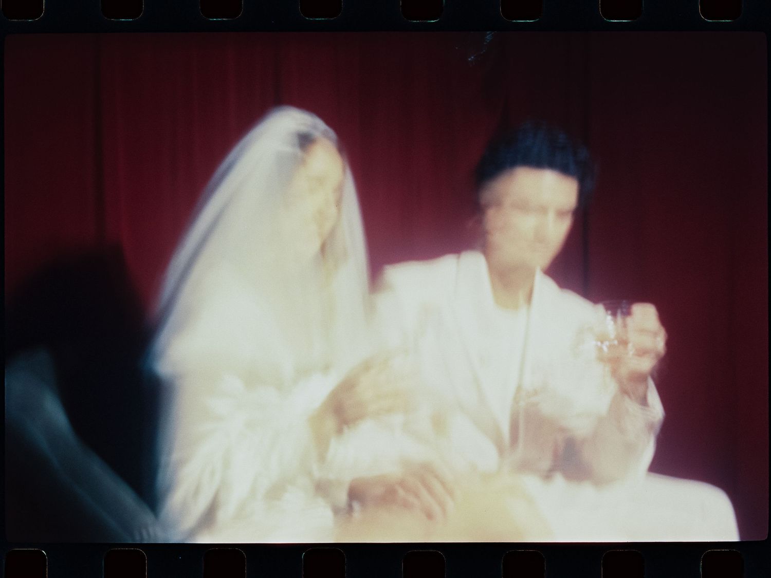 A bride in white veil and groom in white suit sit together against a red curtain backdrop in a vintage film photo.