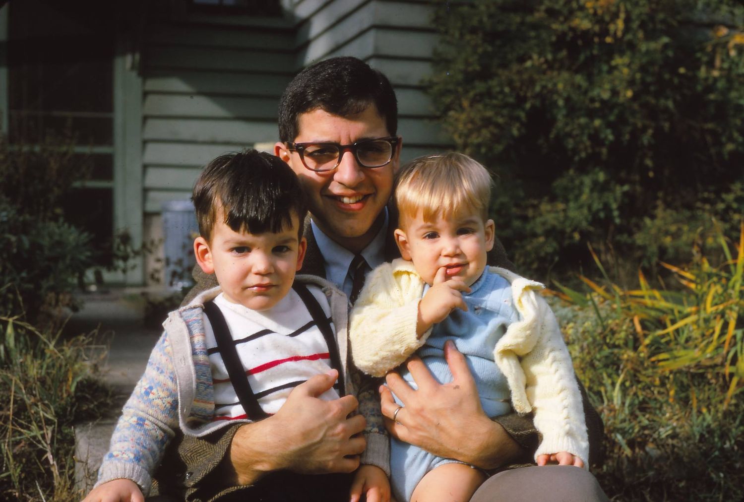 Family group sitting together outdoors on steps in front of house with greenery in background.