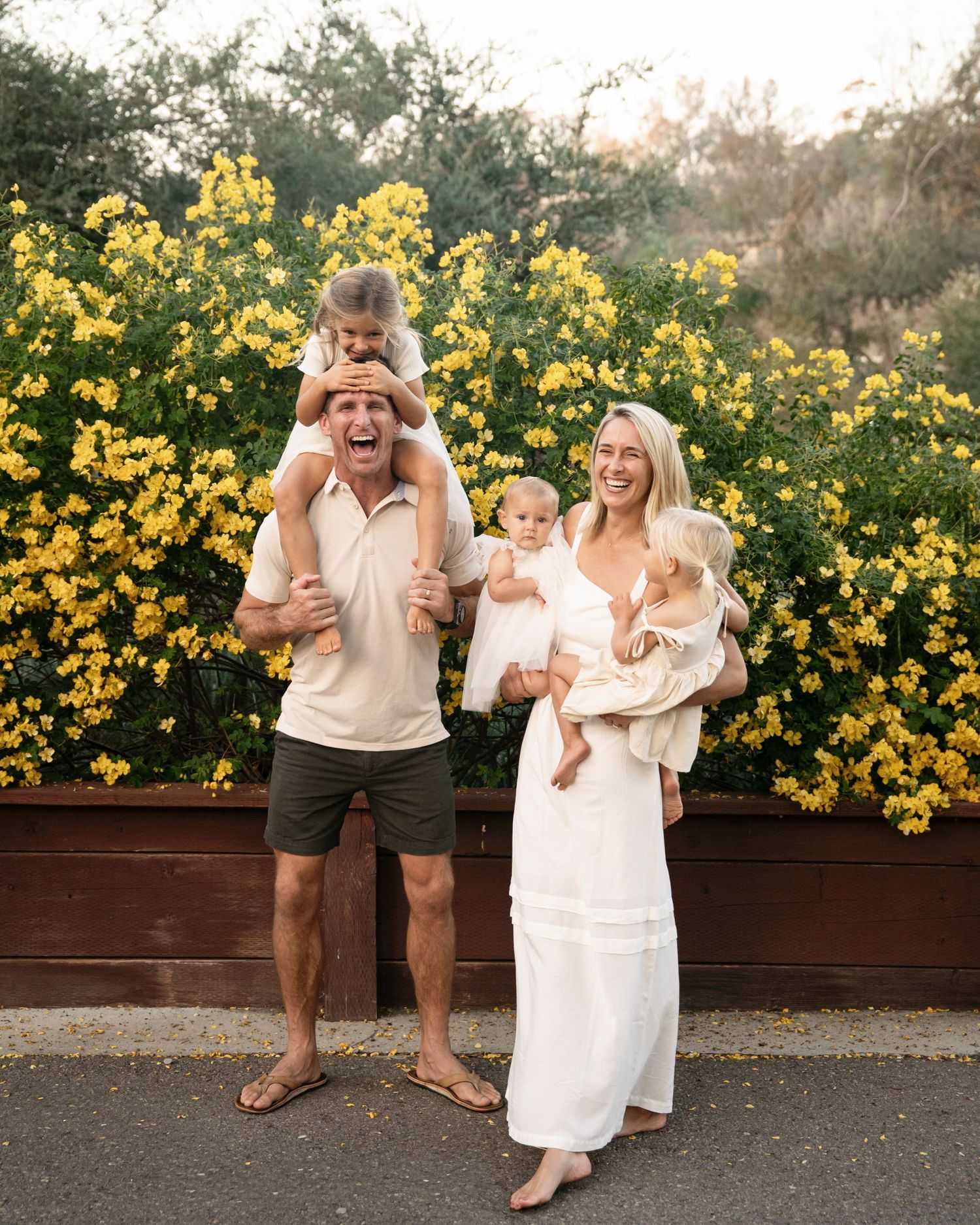 Happy family posing in front of yellow flowering bushes, man with child on shoulders, woman in white dress holding baby.