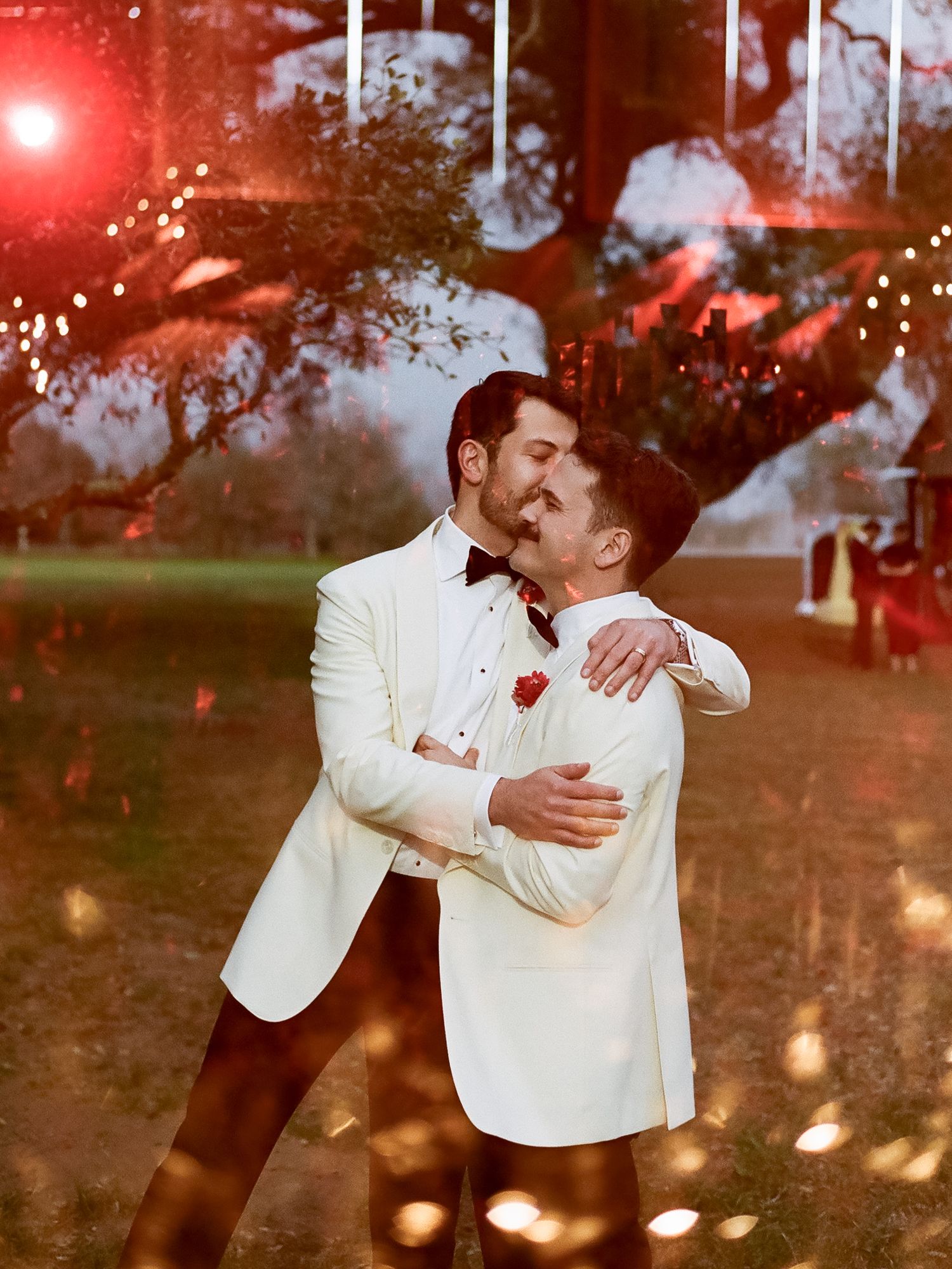 Two grooms in white tuxedos share a romantic embrace outdoors at their evening wedding reception.