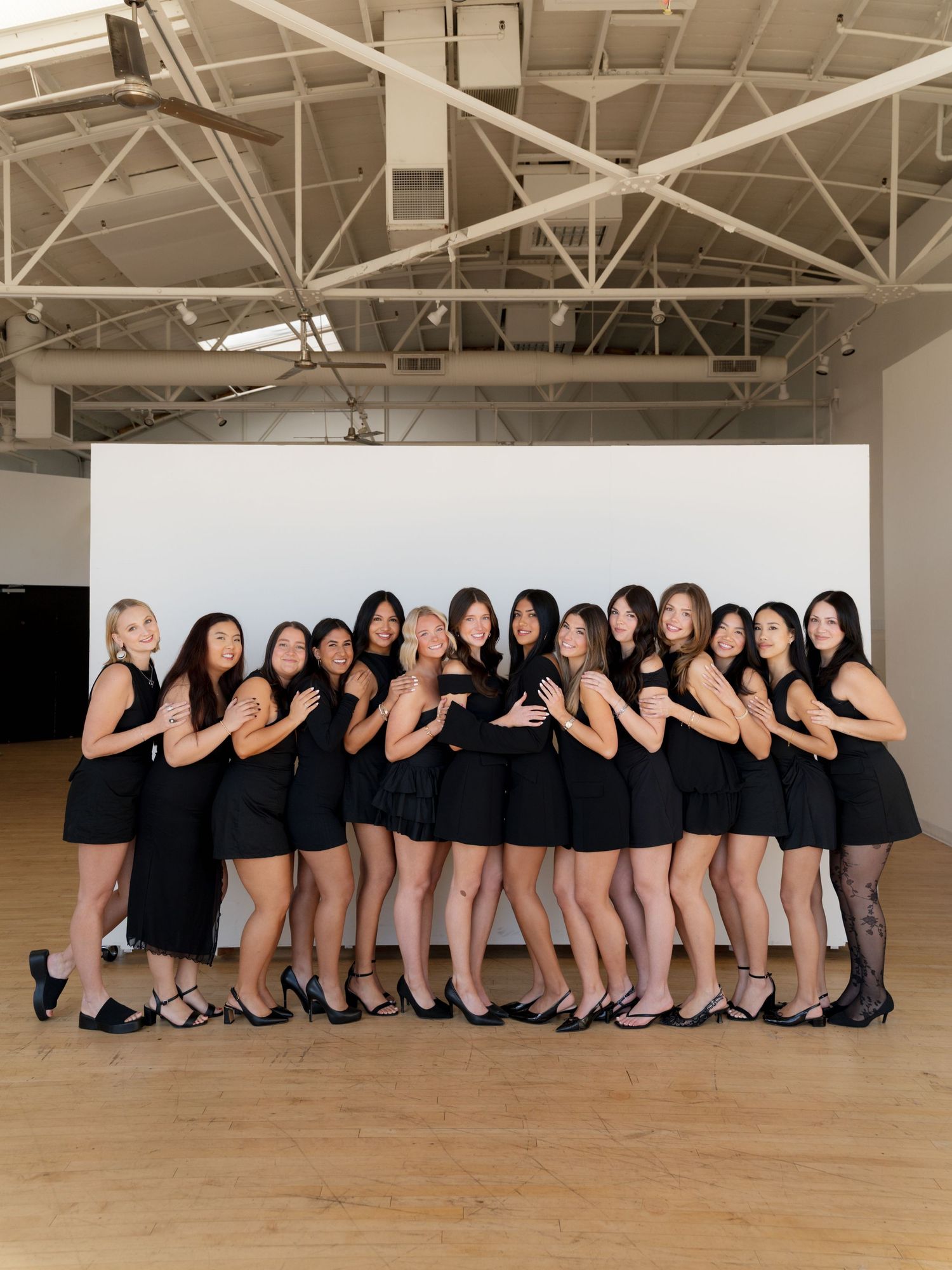 Group of women in matching black dresses posing together in a bright studio space with wooden floors.