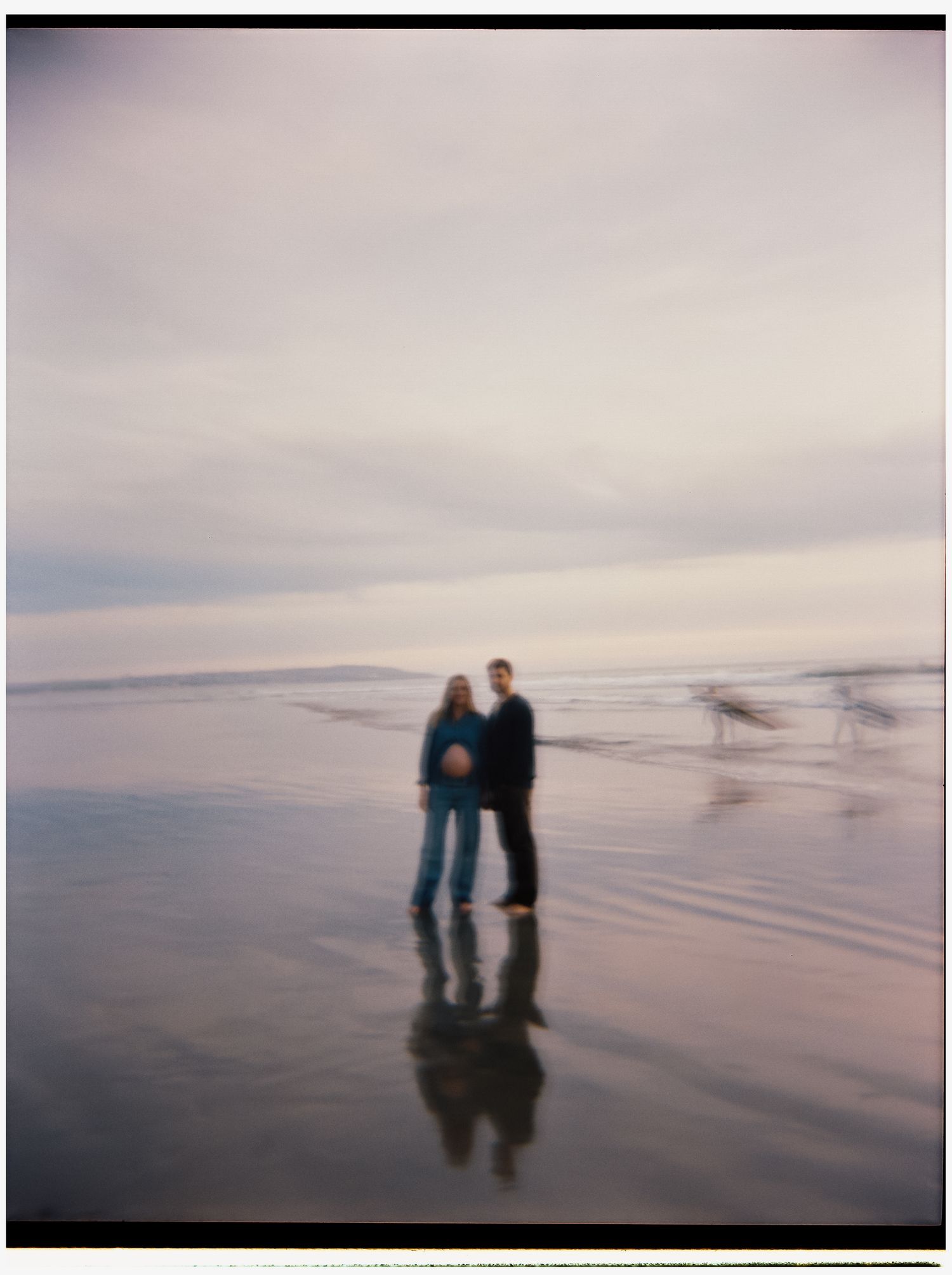 Couple standing together on a reflective beach shore under a cloudy sky, captured in a soft, dreamy film photography style.
