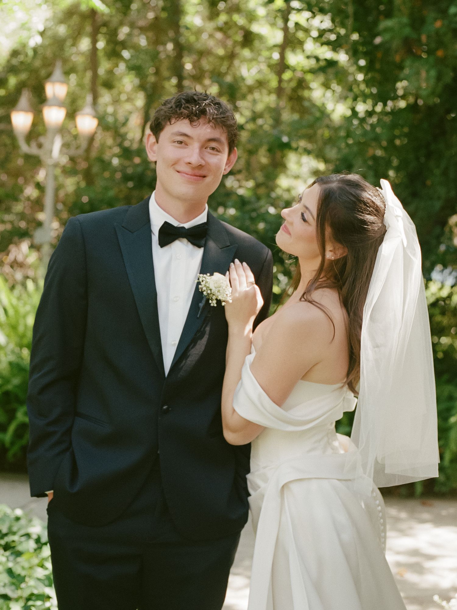 A couple poses in a lush garden, groom in black tuxedo and bride in white off-shoulder gown with veil.