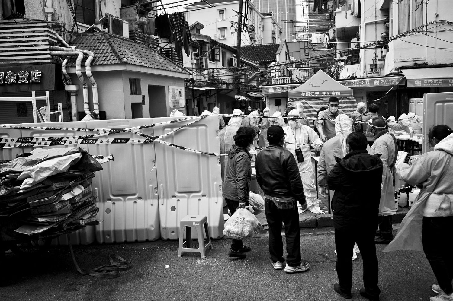 Black and white street photography shows people walking past storefronts and vendor stalls in an urban marketplace.