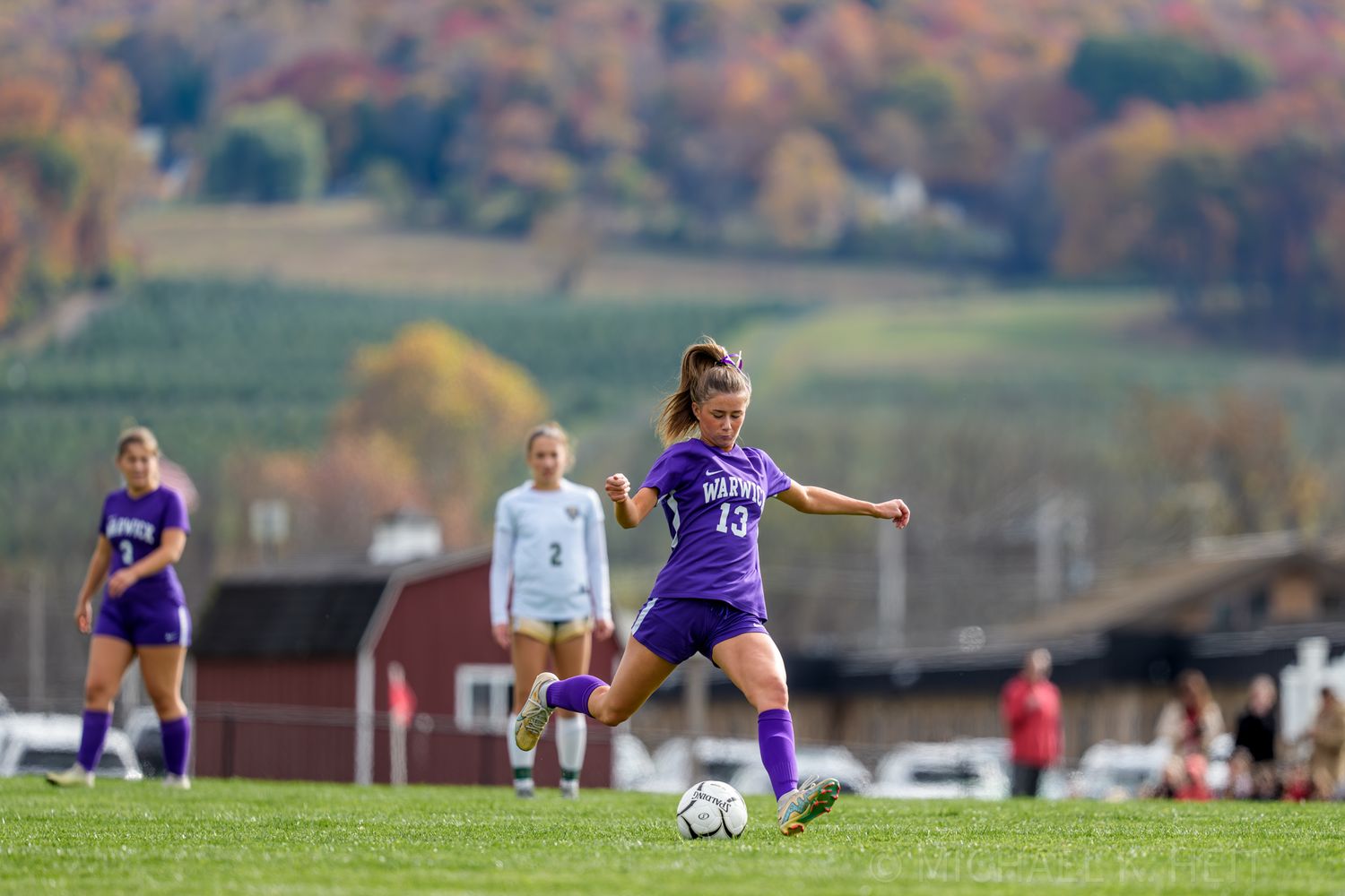 Soccer player in purple jersey takes a powerful kick during a match with autumn foliage in the background.