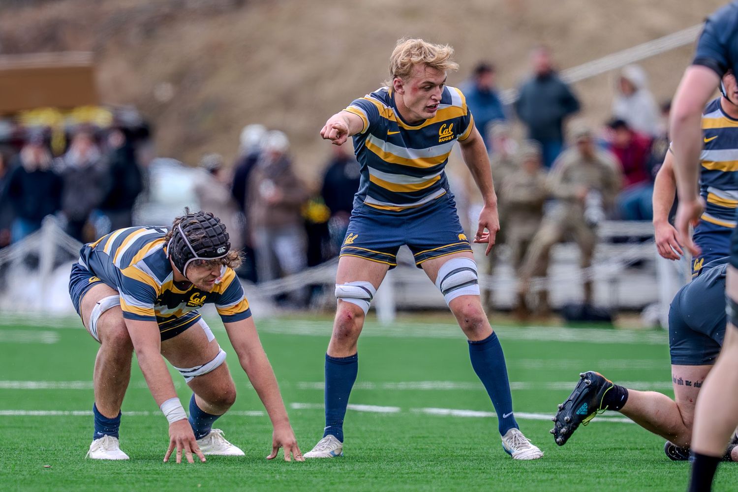 Rugby players in blue and striped uniforms compete for possession during a match on a grass field with spectators.