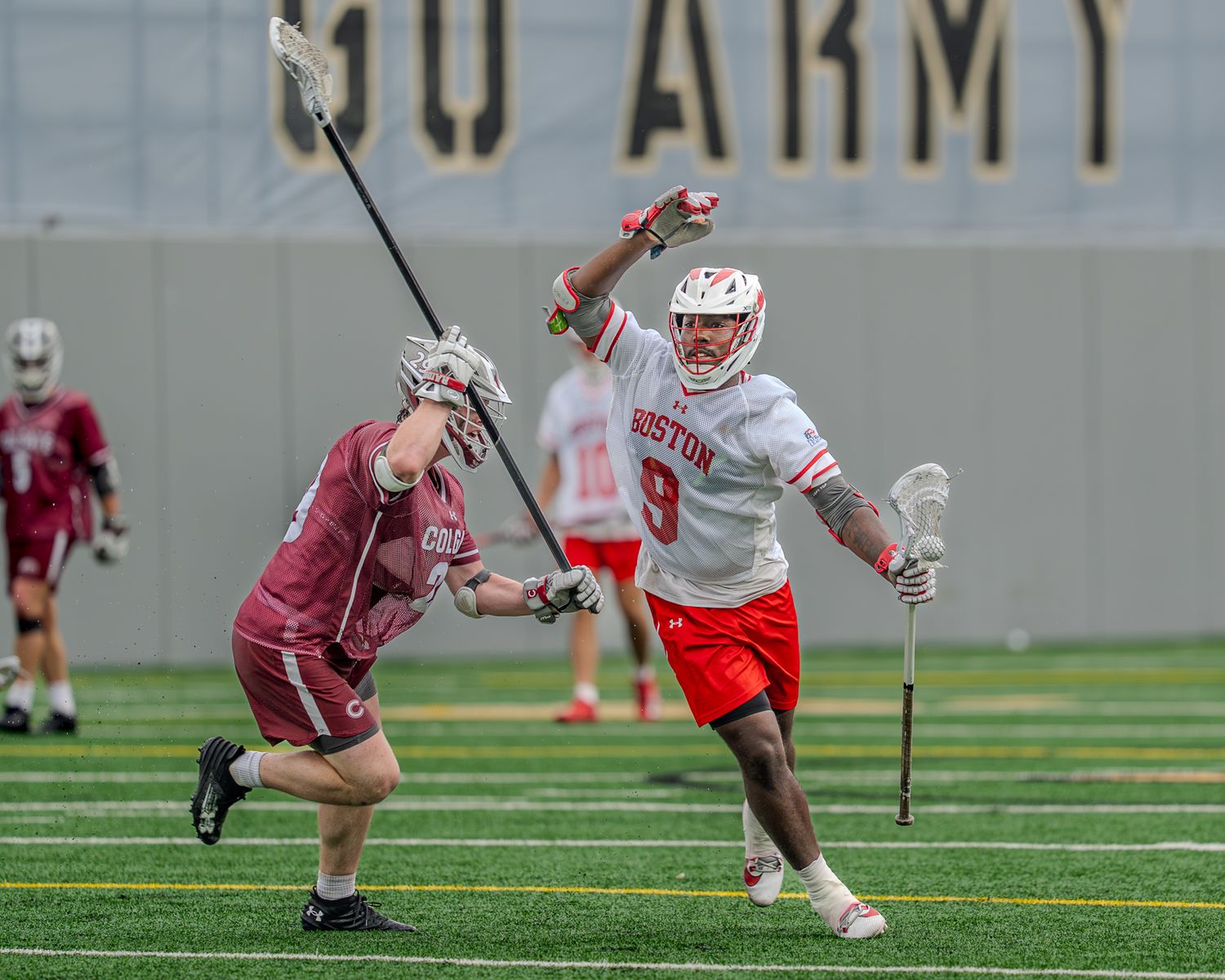 College lacrosse players in red and maroon uniforms compete for possession during an intense match on an artificial turf field.