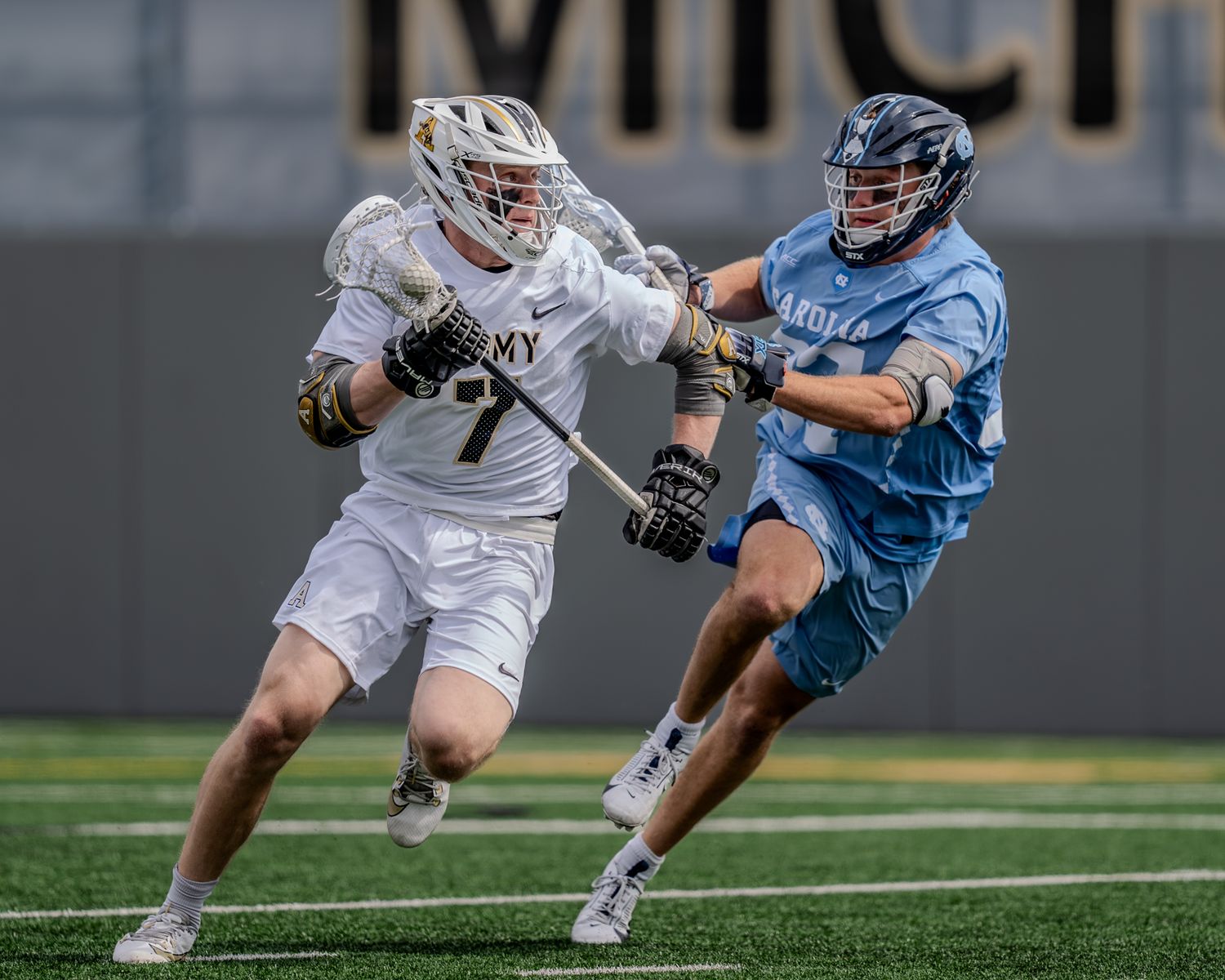 A lacrosse player in white uniform faces off against an opponent in light blue during an intense game moment.