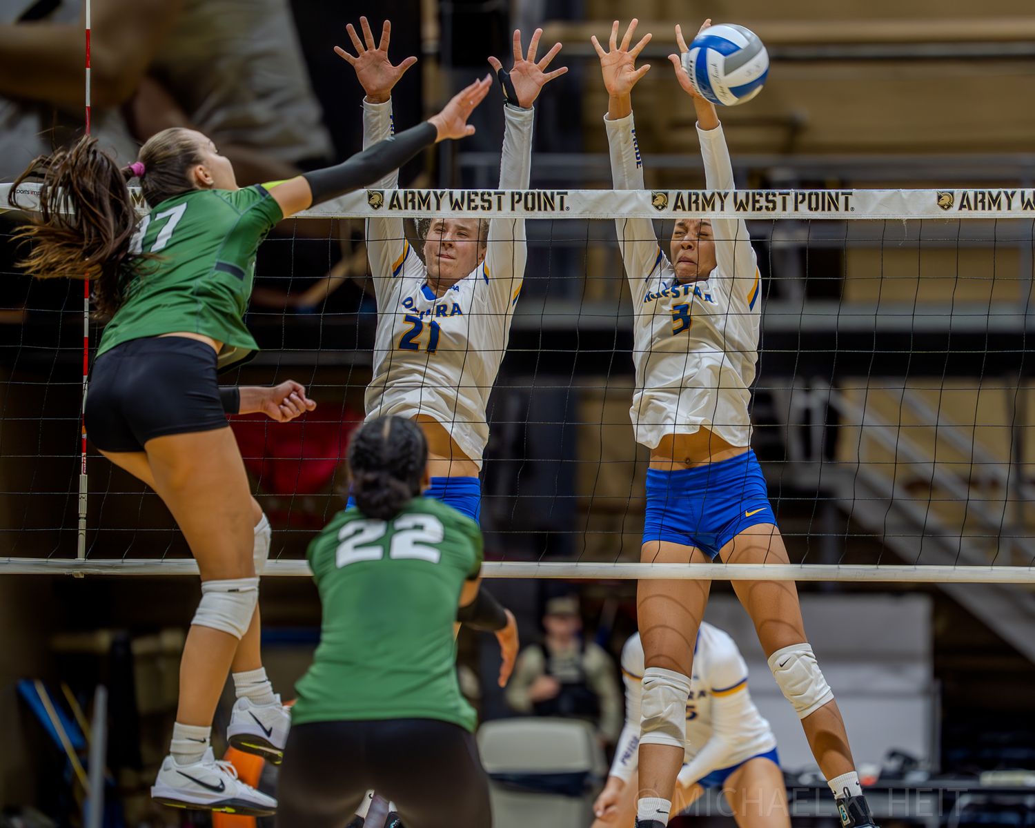 Volleyball players jump high at the net during an intense match between teams in green and white uniforms.