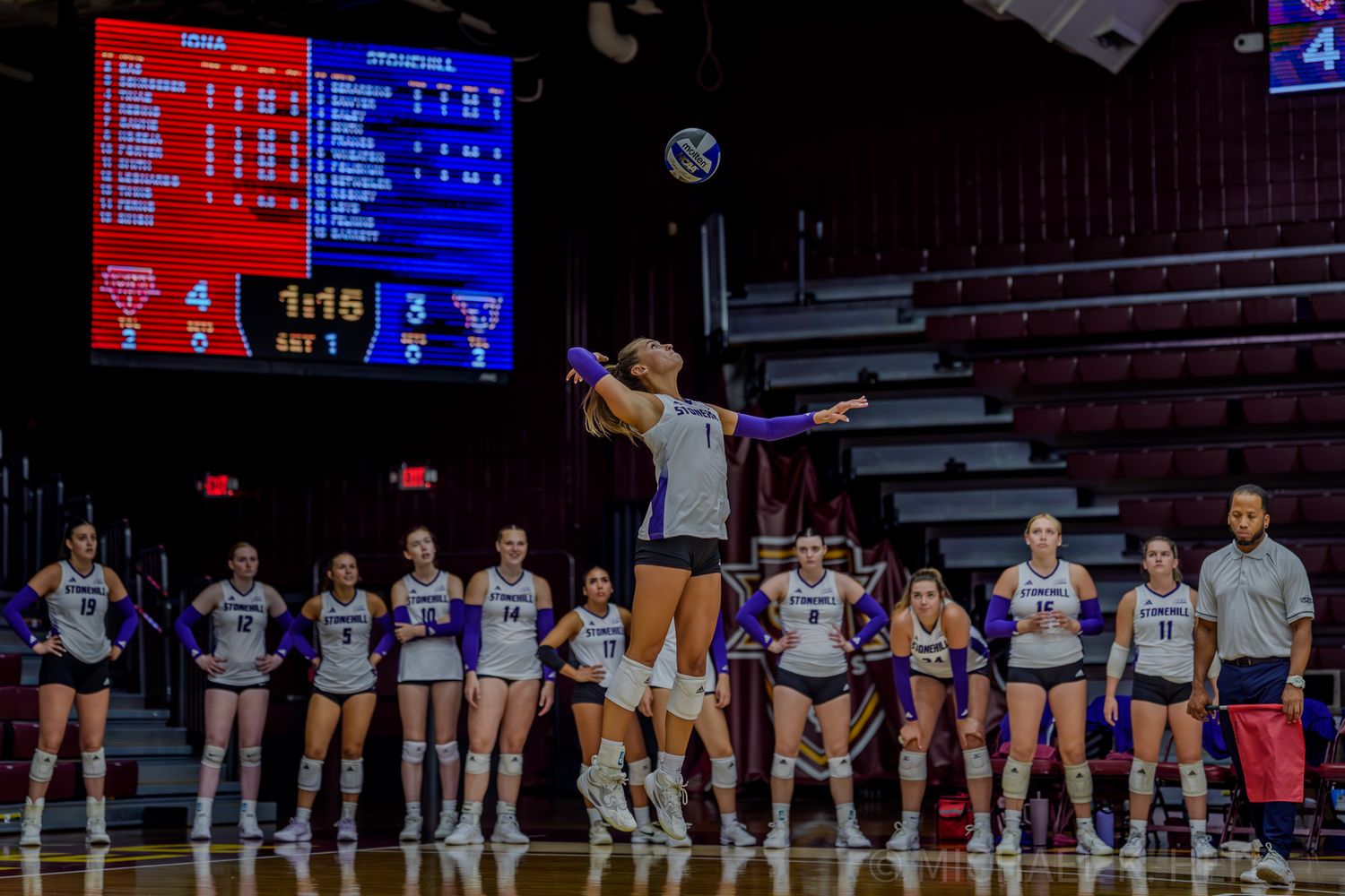 Wide view of volleyball court showing teams lined up during match with electronic scoreboard display.