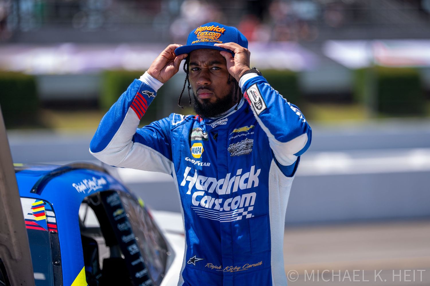 NASCAR driver in Hendrick Cars.com blue racing suit adjusting cap near race car at speedway event.