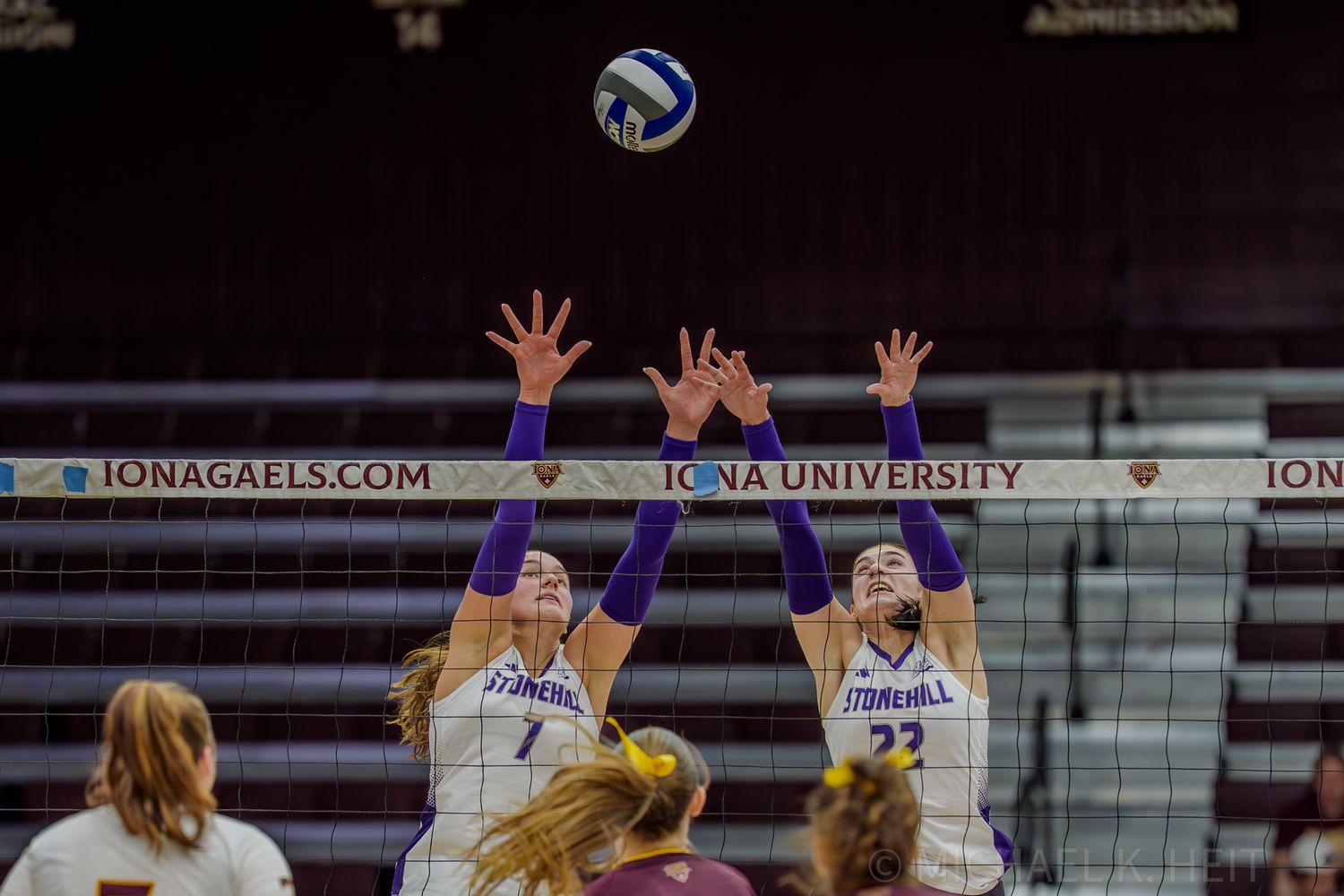 Volleyball players reach high to block a shot during an intense indoor game.