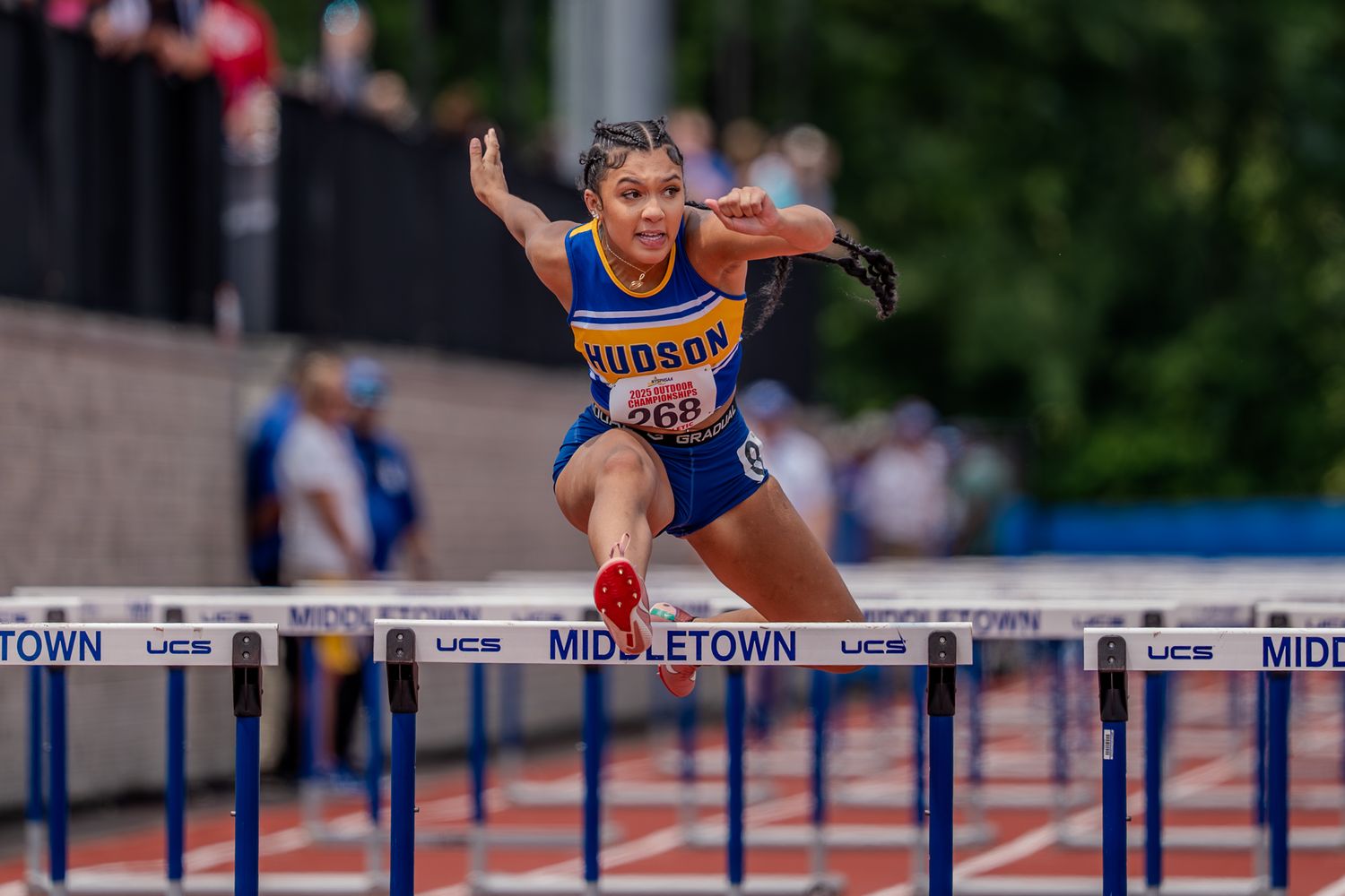 Track athlete clears hurdle during competitive race on outdoor athletics track with multiple lanes.