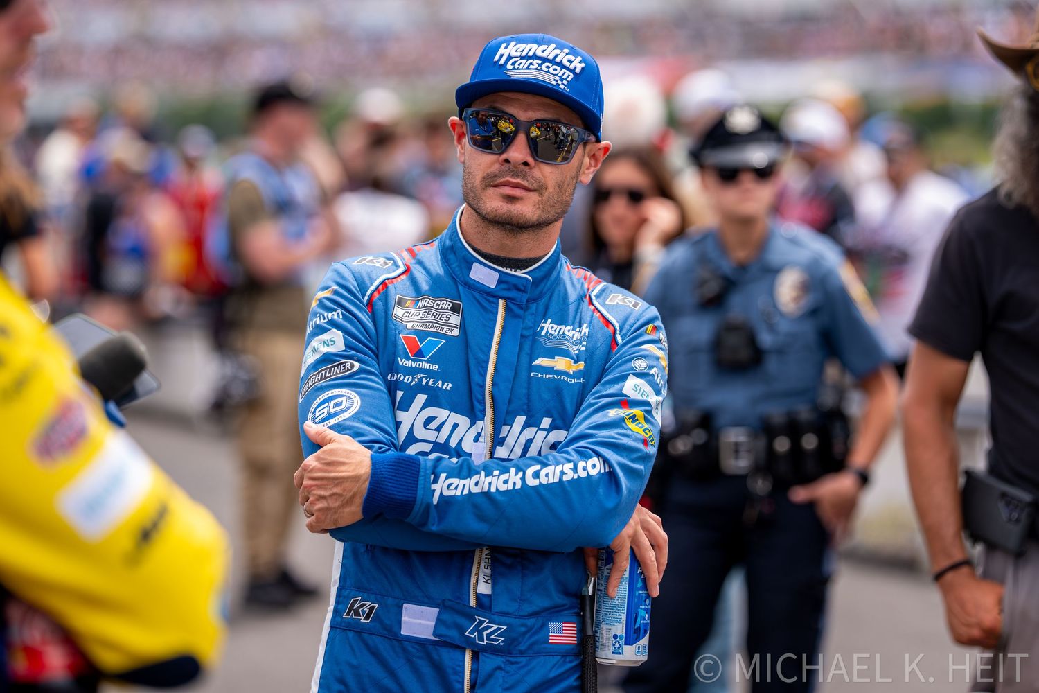 NASCAR driver in blue Hendrick Cars racing suit stands confidently at a race event with police officer in background.