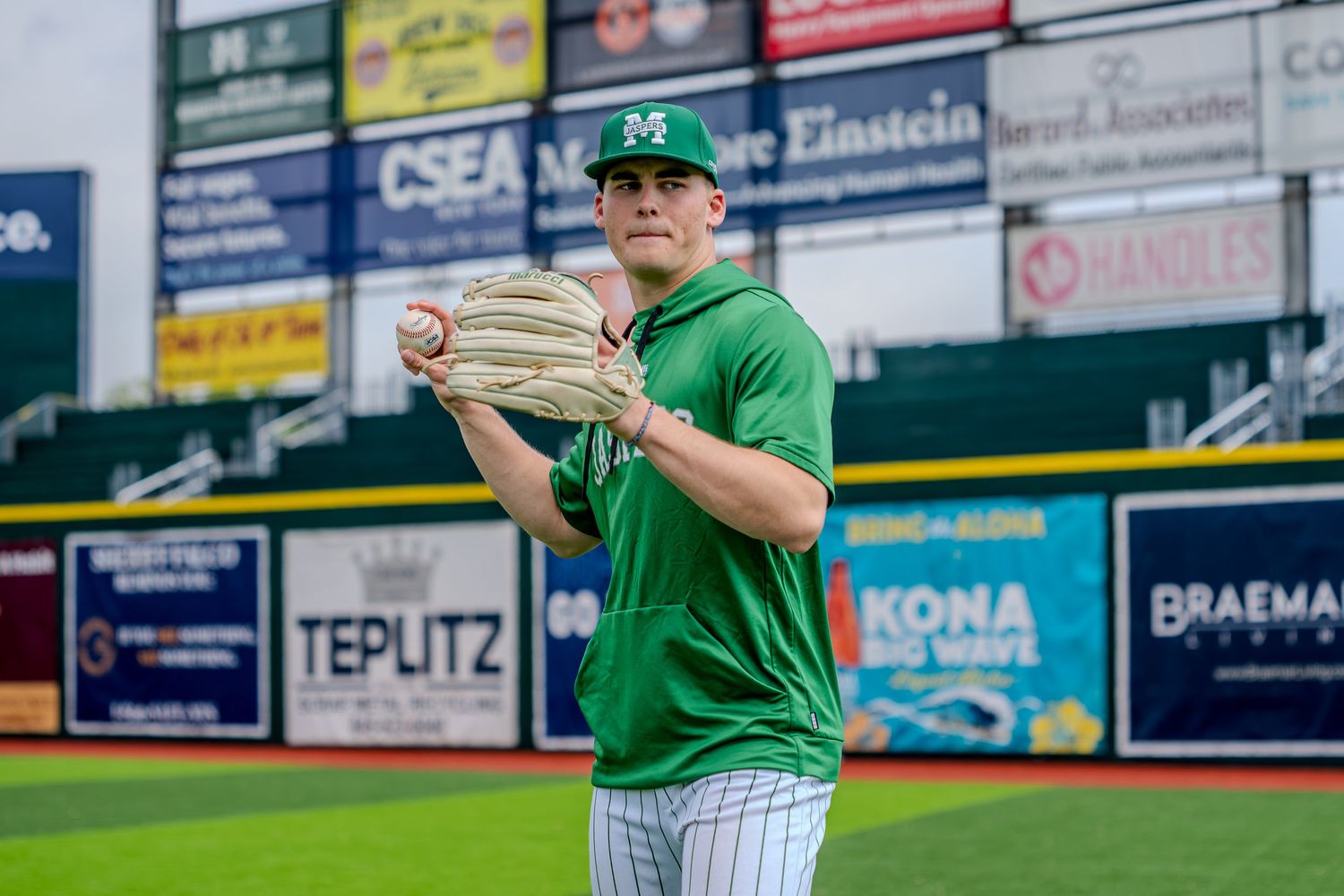 Baseball pitcher in green uniform winds up to throw on a professional baseball field with sponsor banners.