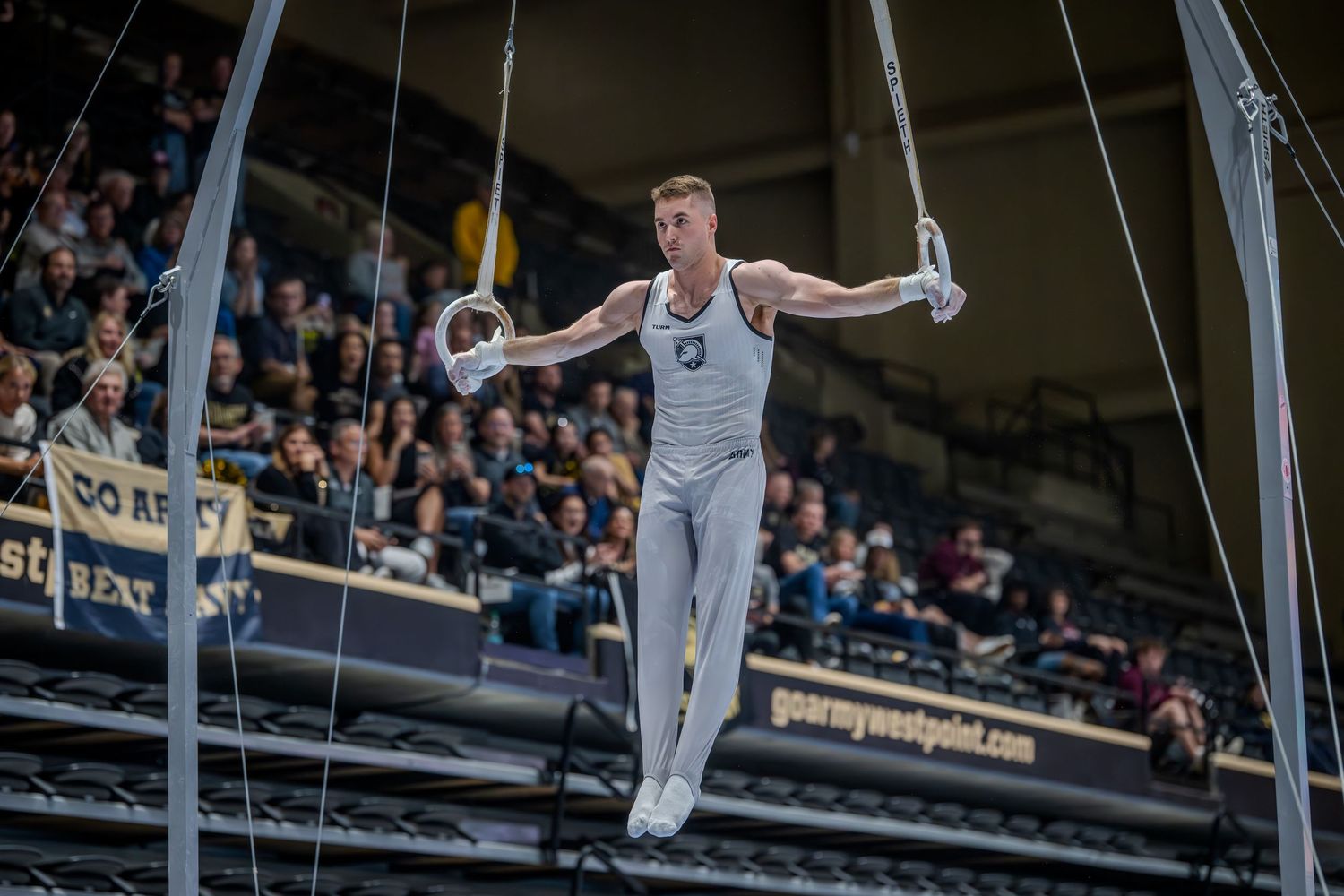Male gymnast performing on still rings at an indoor gymnastics competition with crowd watching in the background.