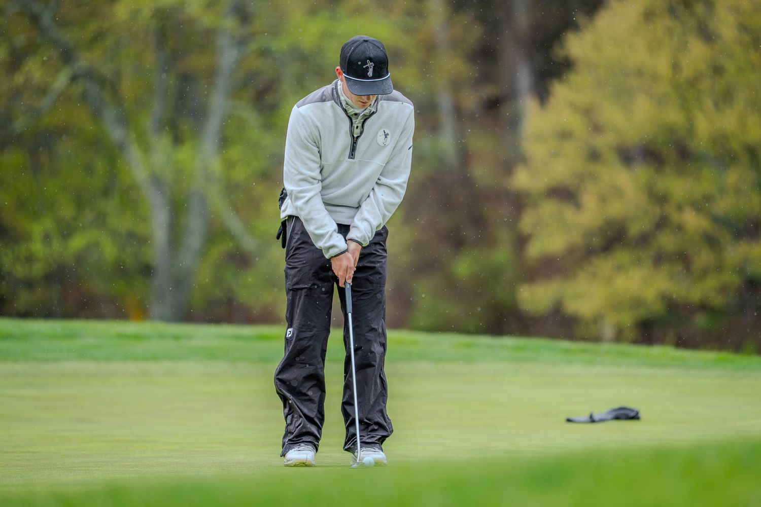Golfer in gray hoodie and black pants focuses on putt on lush green course surrounded by autumn trees.