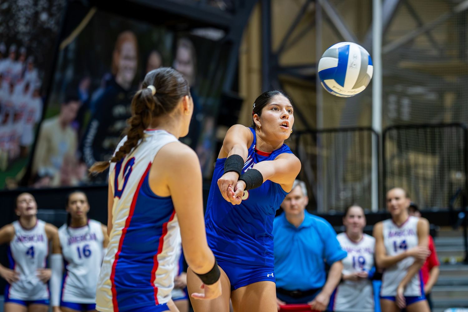 Female volleyball player in blue uniform performs a dig during an indoor collegiate volleyball match.
