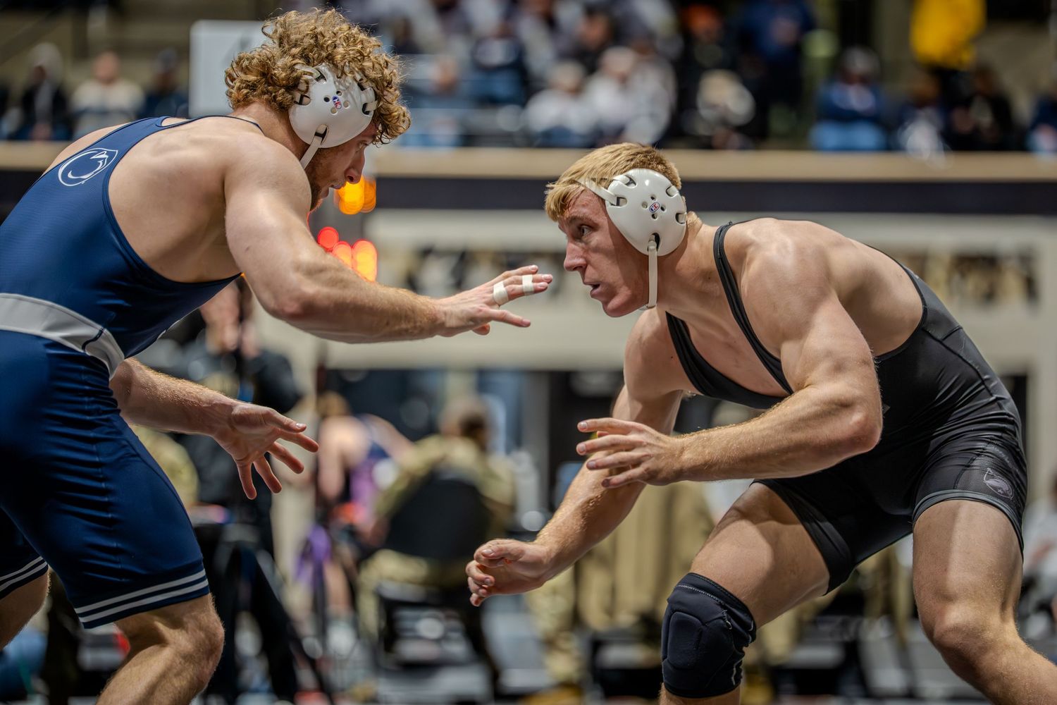 Two wrestlers in headgear compete on a mat, one in blue Penn State singlet, the other in black, in an intense college wrestling match.