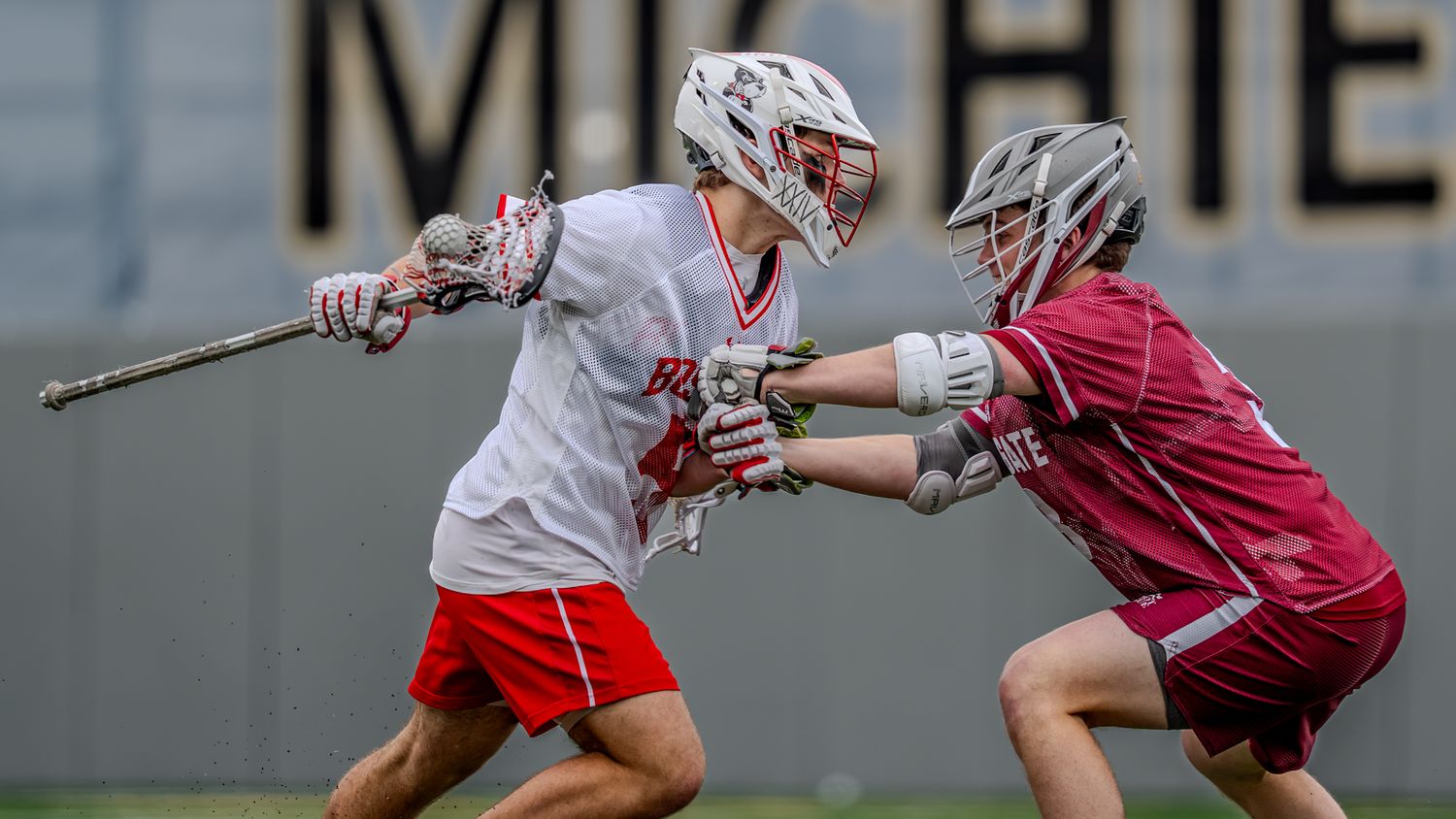 Two lacrosse players in red and white uniforms compete for possession during a match.